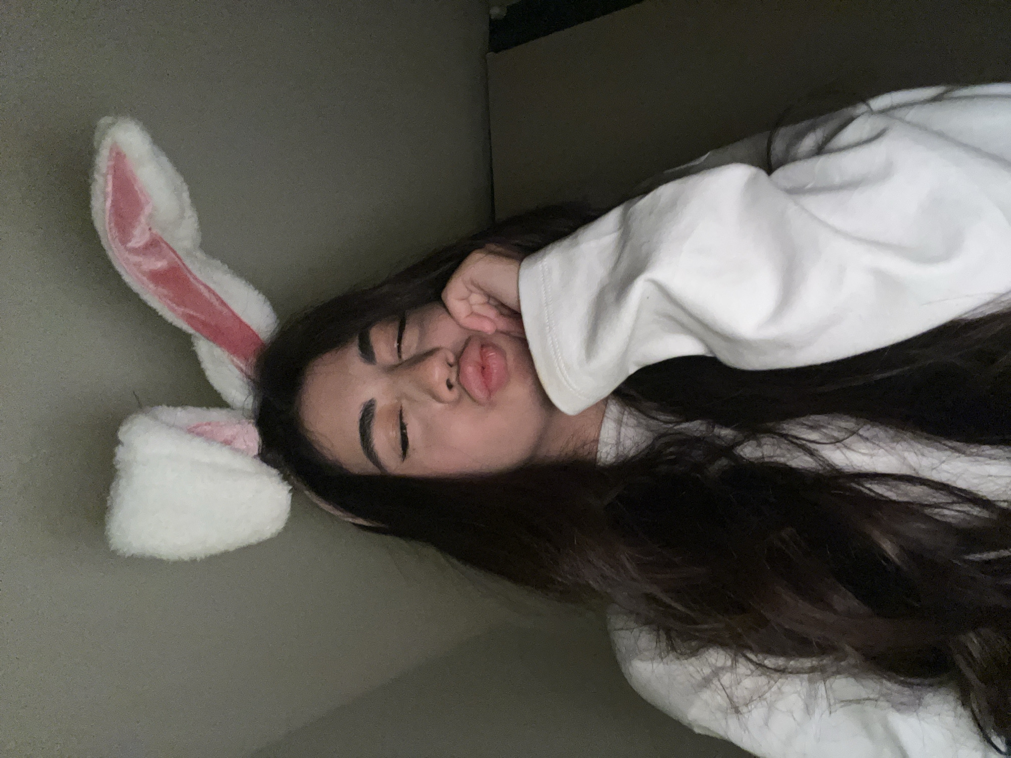 A young woman with long, dark, wavy hair and a white sweatshirt is wearing fuzzy white bunny ears with pink inner lining. She is leaning her head on her hand, with her eyes closed and her lips pursed in a playful expression. The background is a plain, dark wall. The image does not contain any information that would allow for its location to be determined as Chotěšov, Czechia.