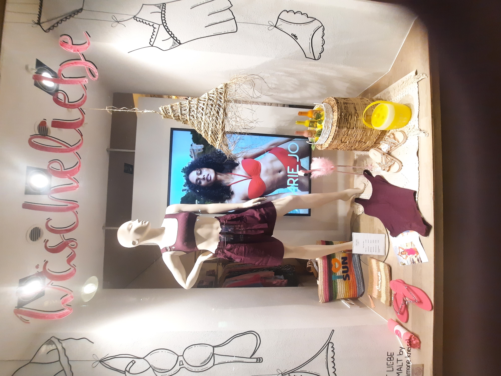 The image shows a retail display window for a lingerie or swimwear store in Rosenheim, Germany. The store's name, "Wäscheliebe", is displayed in pink lettering above the window.
In the center of the display is a female mannequin wearing a maroon bikini top and matching shorts. To the left of the mannequin is a straw bag with colorful stripes and the word "SUN" embroidered on it. In front of the mannequin are various items including pink flip-flops, a pink rolled-up towel, a small clutch purse, a dark maroon swimsuit, and a small photo of a woman in a pink swimsuit.
To the right of the mannequin, a large framed advertisement features a woman in a red bikini. A wicker basket sits on another wicker basket, holding a tray with three bottles and some lemons. A pink flamingo figurine stands to the left of the baskets. A bright yellow cooler is placed on the floor to the right of the basket. The background of the display includes a white wall with black line drawings of bras and other lingerie items. The display appears to be promoting summer or beachwear.