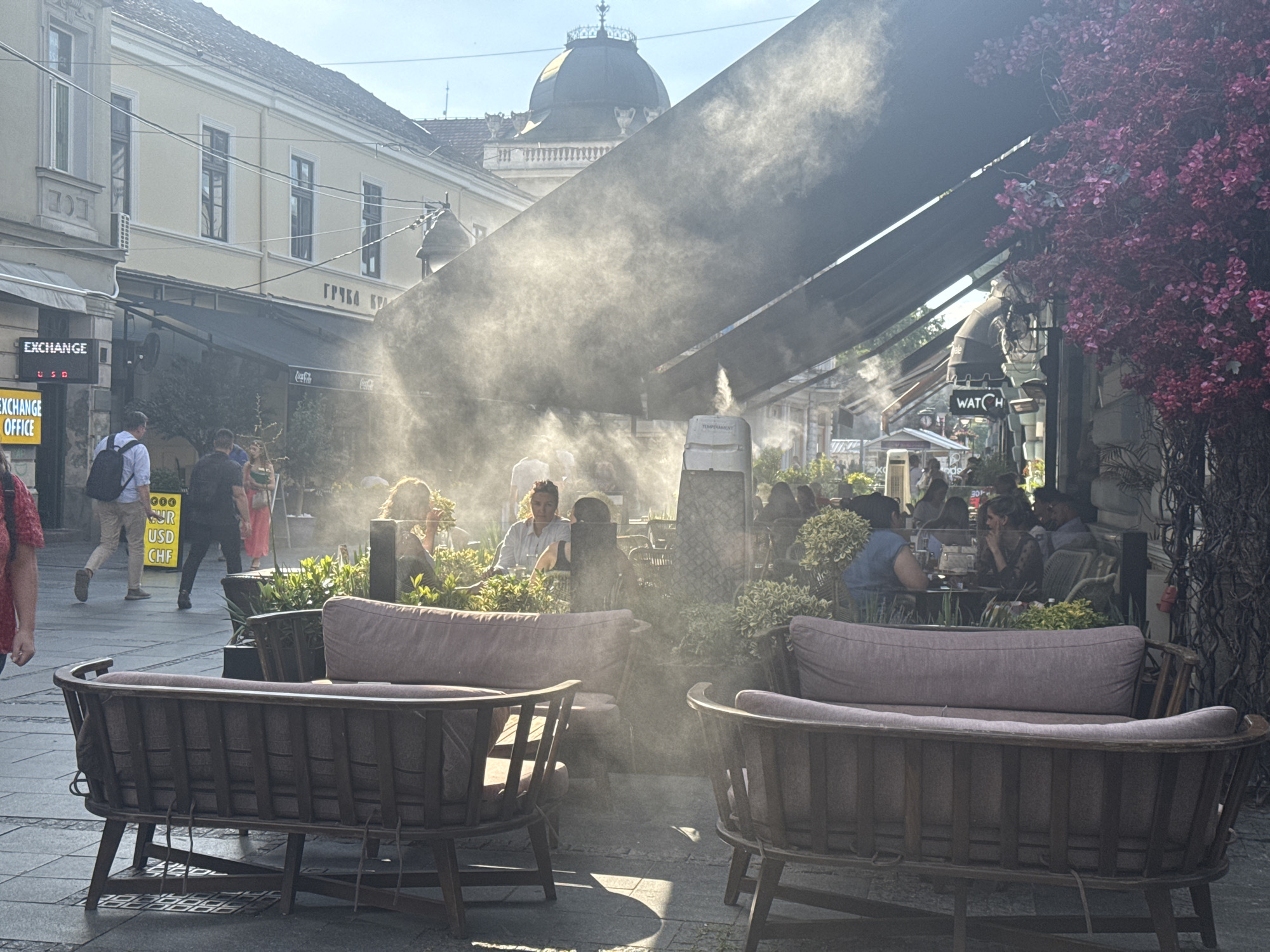 The image depicts a bustling outdoor seating area of a cafe or restaurant on a sunny day in Belgrade, Serbia. The scene is characterized by a fine mist spraying from overhead, creating an ethereal atmosphere and likely serving to cool patrons. Two dark wooden benches with plush mauve cushions are positioned in the foreground, facing away from the camera, suggesting a seating arrangement for customers.
In the midground, several individuals are seated at tables under a large, dark awning, engaged in conversation and enjoying their time. The mist obscures some of the details of their faces and interactions, but their presence indicates a lively social environment. Further back, buildings with classical architectural features, including windows and a domed structure, are visible against a clear blue sky, providing context for the urban setting.
Signs for "EXCHANGE OFFICE" and "WATCH" are visible on some of the buildings, hinting at the commercial nature of the area. Lush green foliage and a vibrant pink flowering plant add a touch of natural beauty to the scene, contrasting with the urban landscape. The overall impression is one of a pleasant, social gathering spot in Belgrade, enhanced by the cooling mist and the warm sunlight.