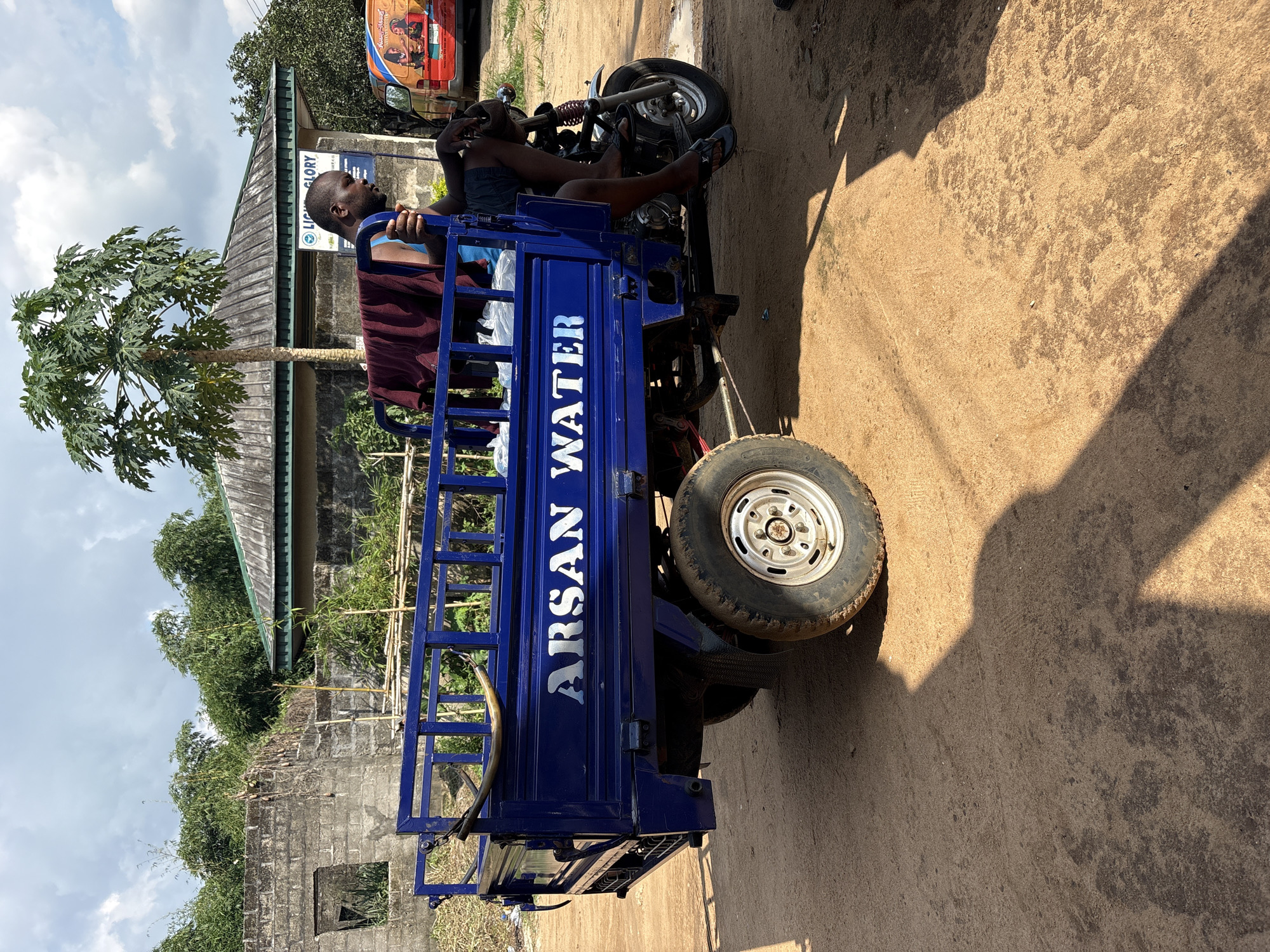 A man sits on a blue three-wheeled motorcycle with "ARSAN WATER" printed on the side, parked on a dirt road. The backdrop includes a building with a sign that reads "LIFE GLORY." In the background is Ikot Ekpene, Nigeria, where the scene takes place.