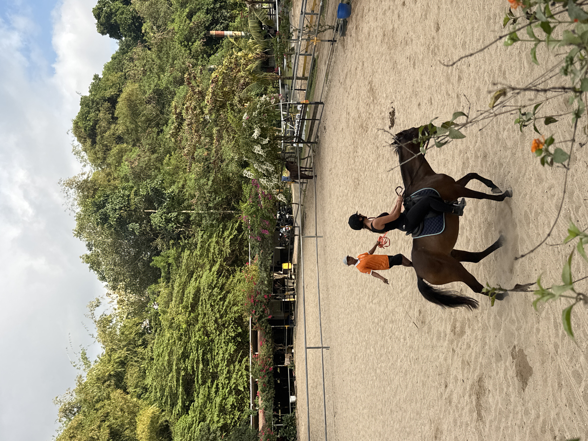 In Kuta Selatan, Indonesia, a horseback riding arena is depicted. The sandy arena is surrounded by lush green foliage and trees. In the foreground, a woman wearing a black riding helmet and outfit is mounted on a brown horse. She is holding the reins and appears to be in motion. To her left and slightly behind, a man in an orange shirt and black shorts is walking and holding a red rope or lead. He seems to be guiding or assisting the rider and horse. The background shows a fenced-off area with stables and more trees. The sky is partly cloudy with patches of blue. The overall scene suggests an outdoor equestrian activity in a tropical environment.