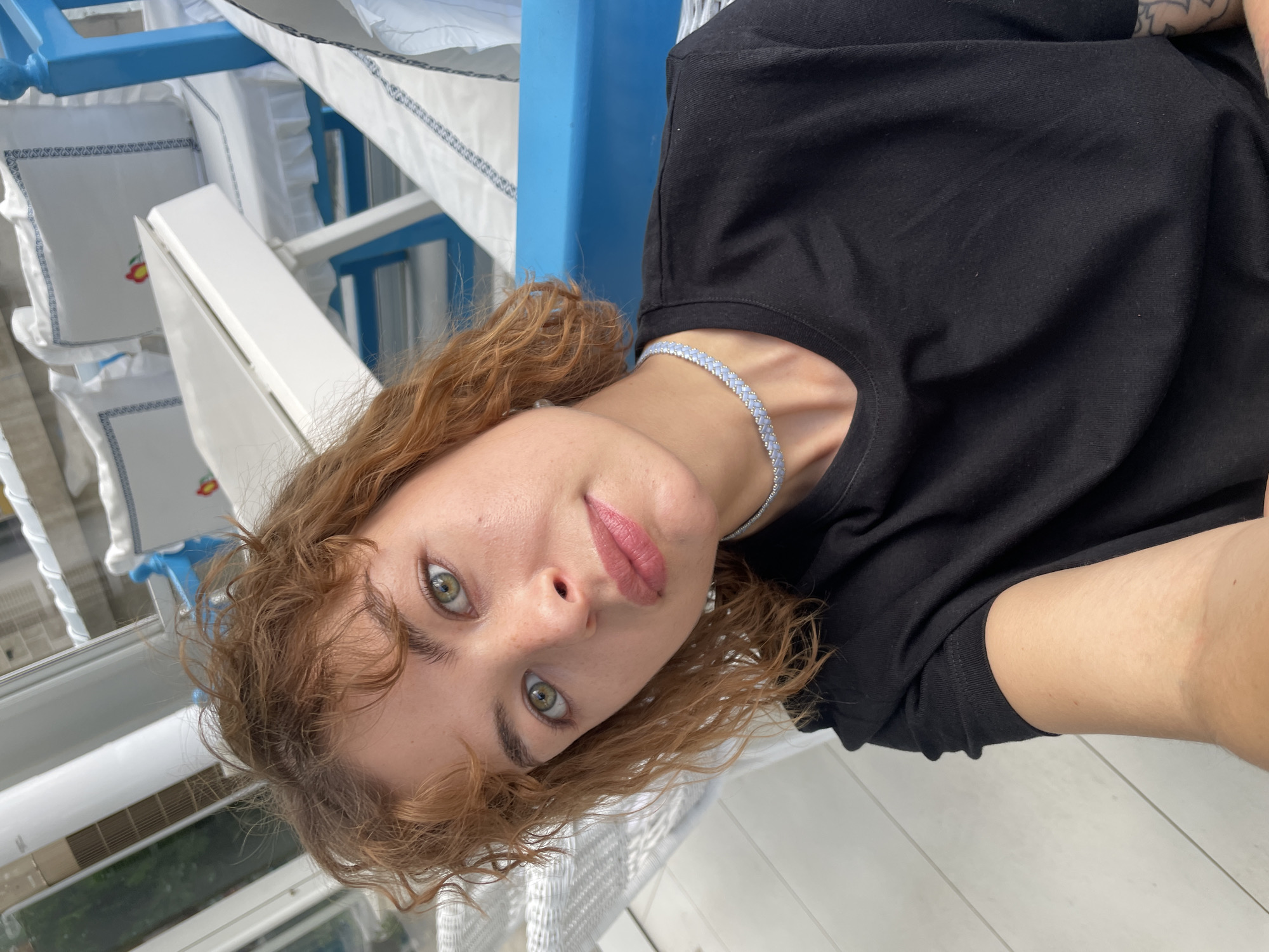 A woman with curly reddish-brown hair and green eyes is wearing a black t-shirt and a blue and silver choker necklace. She is positioned close to the camera in a selfie-style shot. The background features a blue and white structure, potentially part of a boat or a building. Small white decorative pillows with embroidered cherries can be seen in the background. The location is likely Podgorica, Montenegro, given the architectural details.