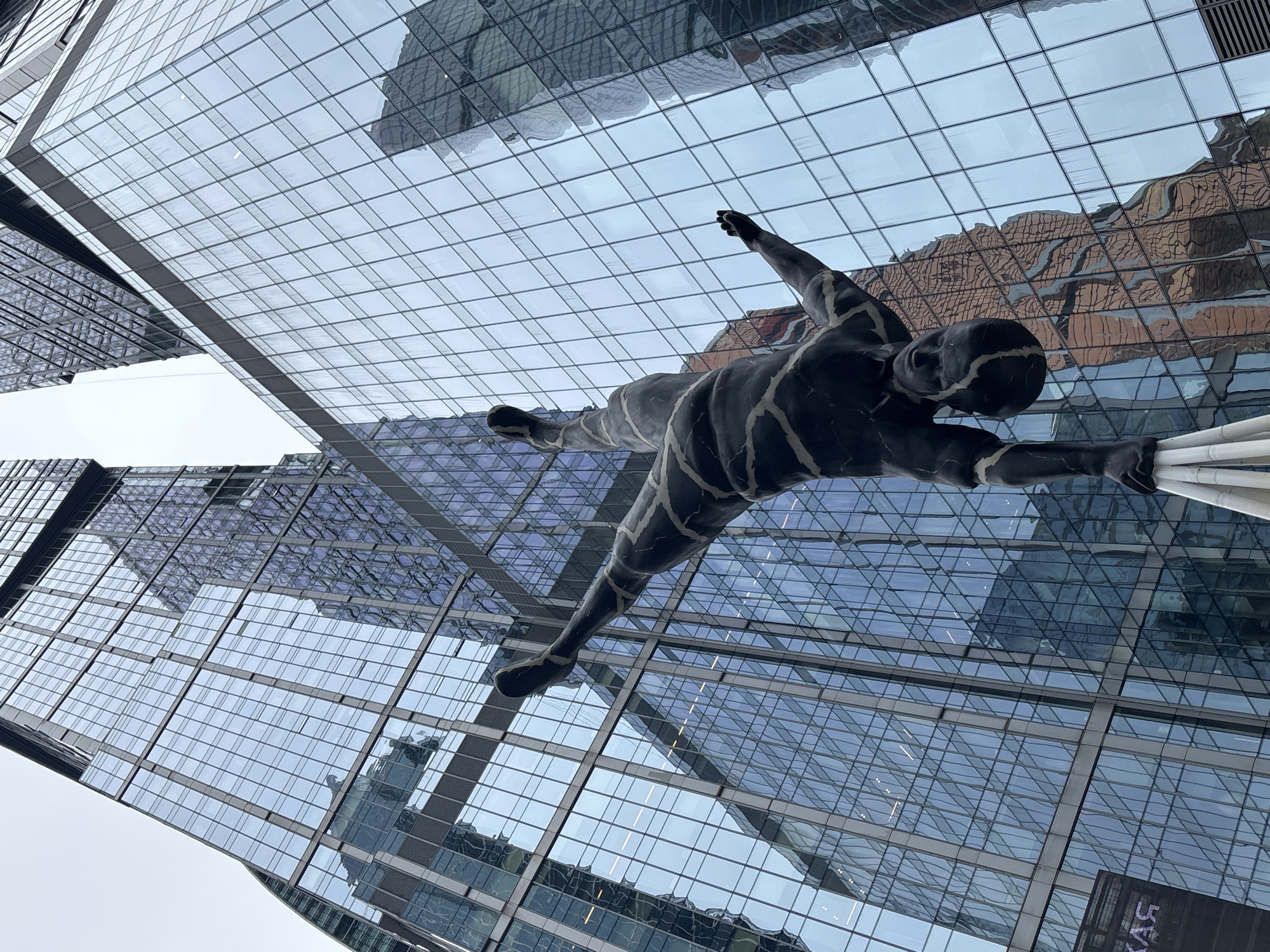 The image shows a low-angle view of the "Thinker" sculpture by Franz Josef Domscheit, located in the Moscow International Business Center (Moscow City), Russia. The sculpture is a dark, patterned figure suspended against a backdrop of modern glass skyscrapers and a cloudy sky. The architecture of the surrounding buildings is reflected in the glass panels. The sculpture appears to be in a diving or reaching pose.