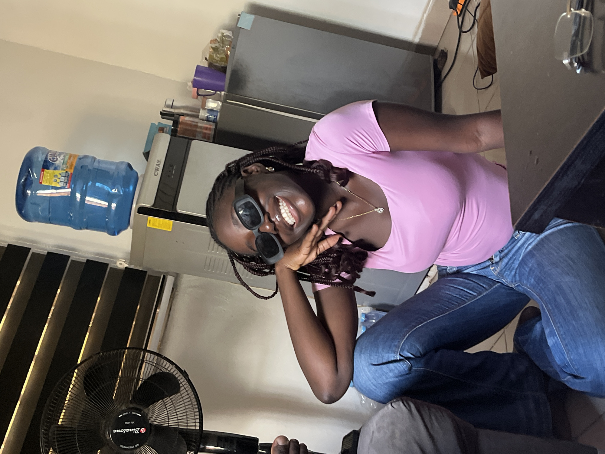 A young, dark-skinned woman is kneeling and smiling at the camera. She is wearing a pink top, blue jeans, and dark sunglasses. Her hair is styled in braids. She has a gold necklace. Behind her is a water dispenser with a large bottle of water on top, and a stainless-steel refrigerator. A black floor fan is to her left. The location appears to be indoors in Abuja, Nigeria. Other people are partially visible in the foreground.