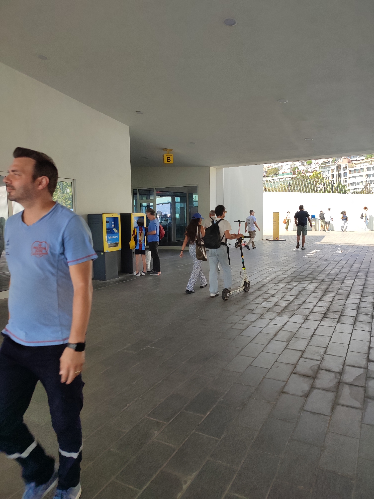 The image depicts an outdoor scene, likely a public walkway or promenade, with a modern architectural overhang. In the foreground, a man wearing a blue t-shirt with red trim and dark pants is walking towards the left of the frame. He has dark hair and appears to be in his late 30s or early 40s. To the right of the foreground figure, there are two yellow ticket vending machines. Further back, several people are walking and interacting. One individual is pushing a scooter. In the background, buildings are visible, suggesting an urban environment. The ground is paved with rectangular tiles. A yellow sign with the letter "B" is visible on the overhang. The scene appears to be in a public area with foot traffic. The context provided, that this is in Beyoğlu, Turkey, suggests a busy urban location within that district.