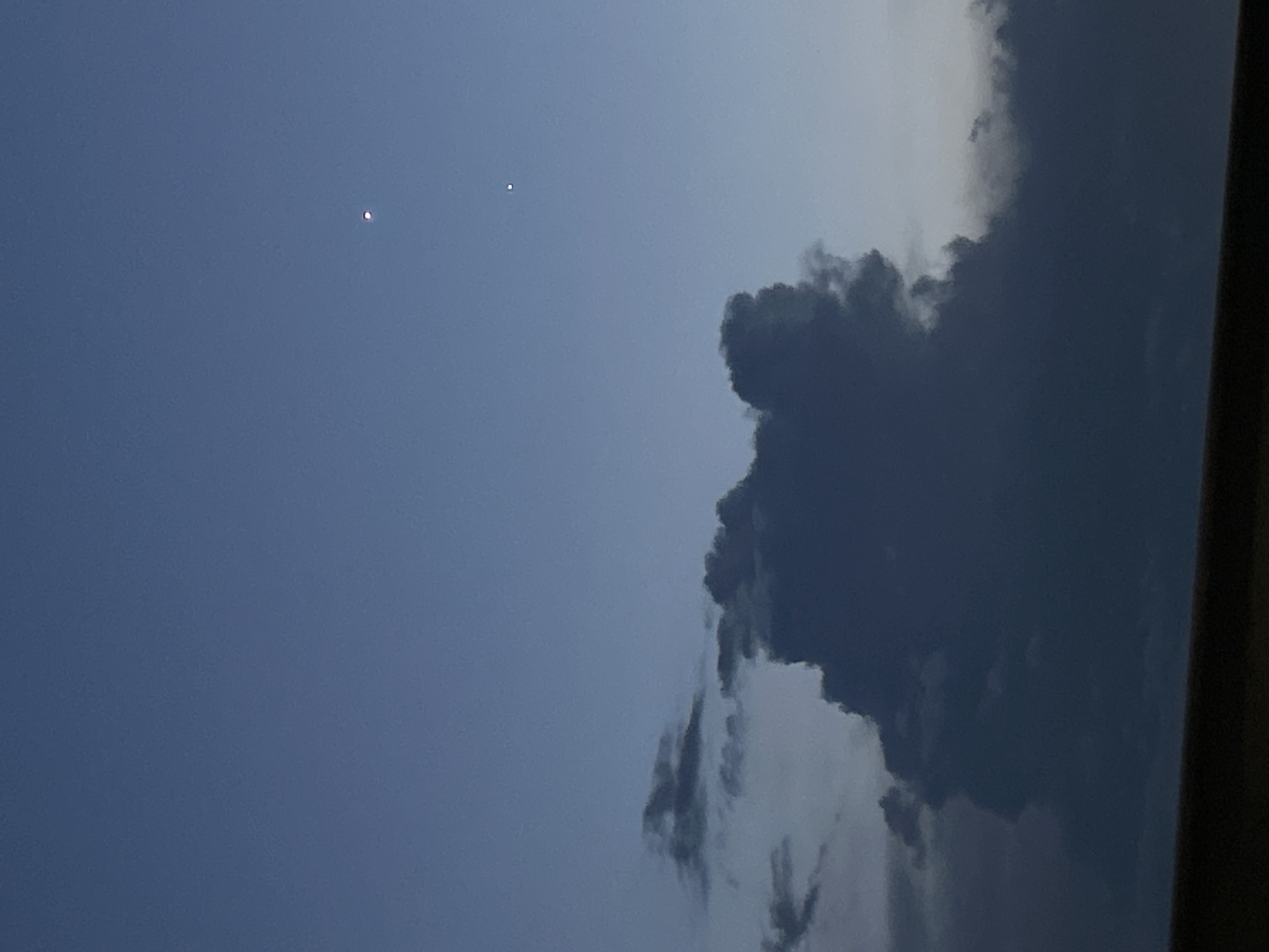 The image shows a dusky sky with a large, dark cloud formation dominating the lower half. Two faint celestial bodies are visible in the upper right quadrant of the sky. The bottom edge of the image features a dark, horizontal band, likely representing the ground or a distant landscape. The scene is set during twilight, indicated by the deep blue hue of the sky and the presence of clouds and celestial bodies. There are no people, animals, buildings, or any specific urban features visible to confirm a location in Sofia, Bulgaria.