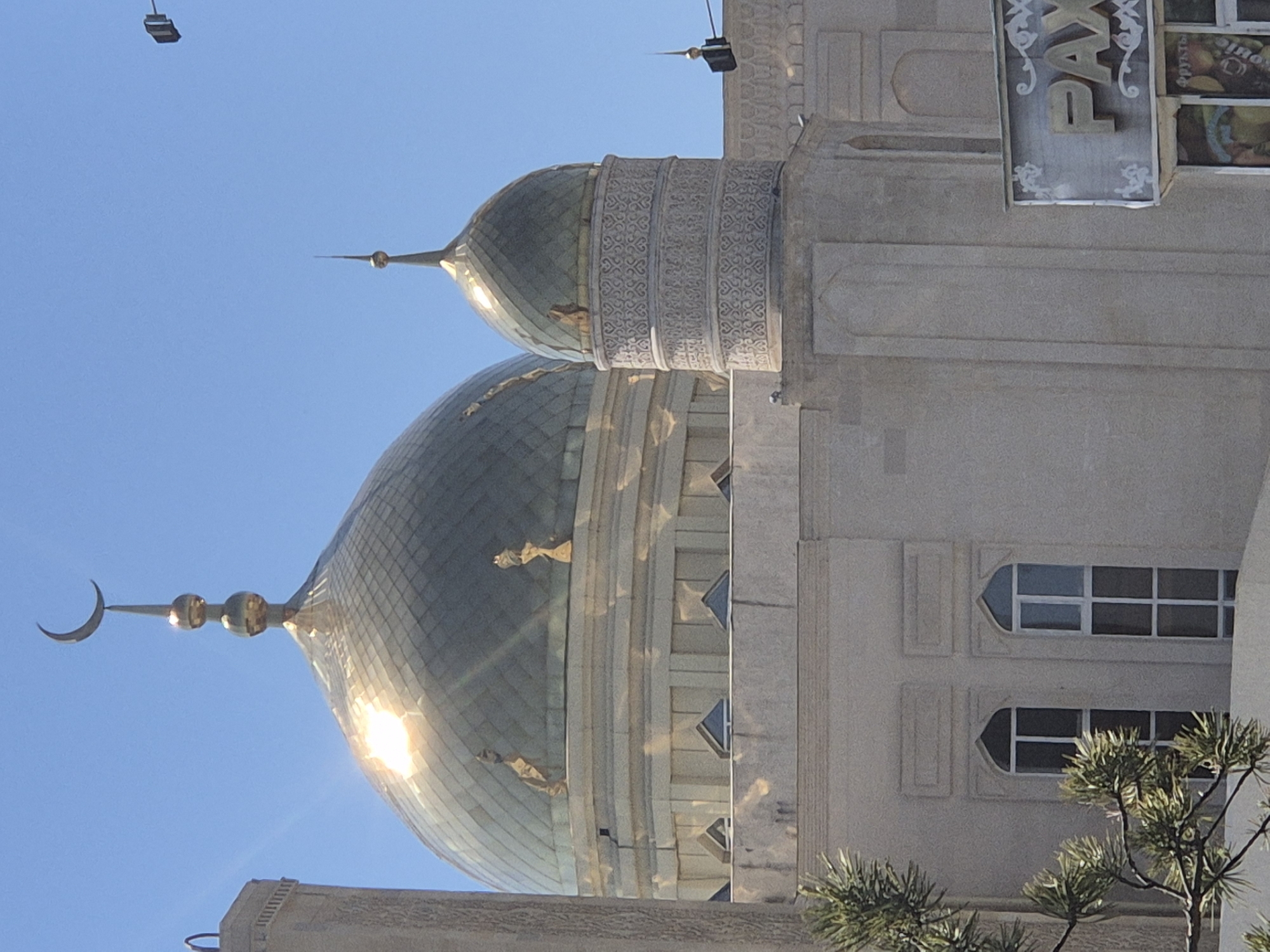 The image shows a section of the Nauryz Mosque in Kokshetau, Kazakhstan. Two of the mosque's golden domes are prominent against a clear blue sky. A crescent moon finial adorns the larger dome. The mosque's structure is light-colored with ornate detailing. A partially visible sign with Cyrillic lettering hangs on the side of the building. A small tree is visible at the bottom right corner of the image.
