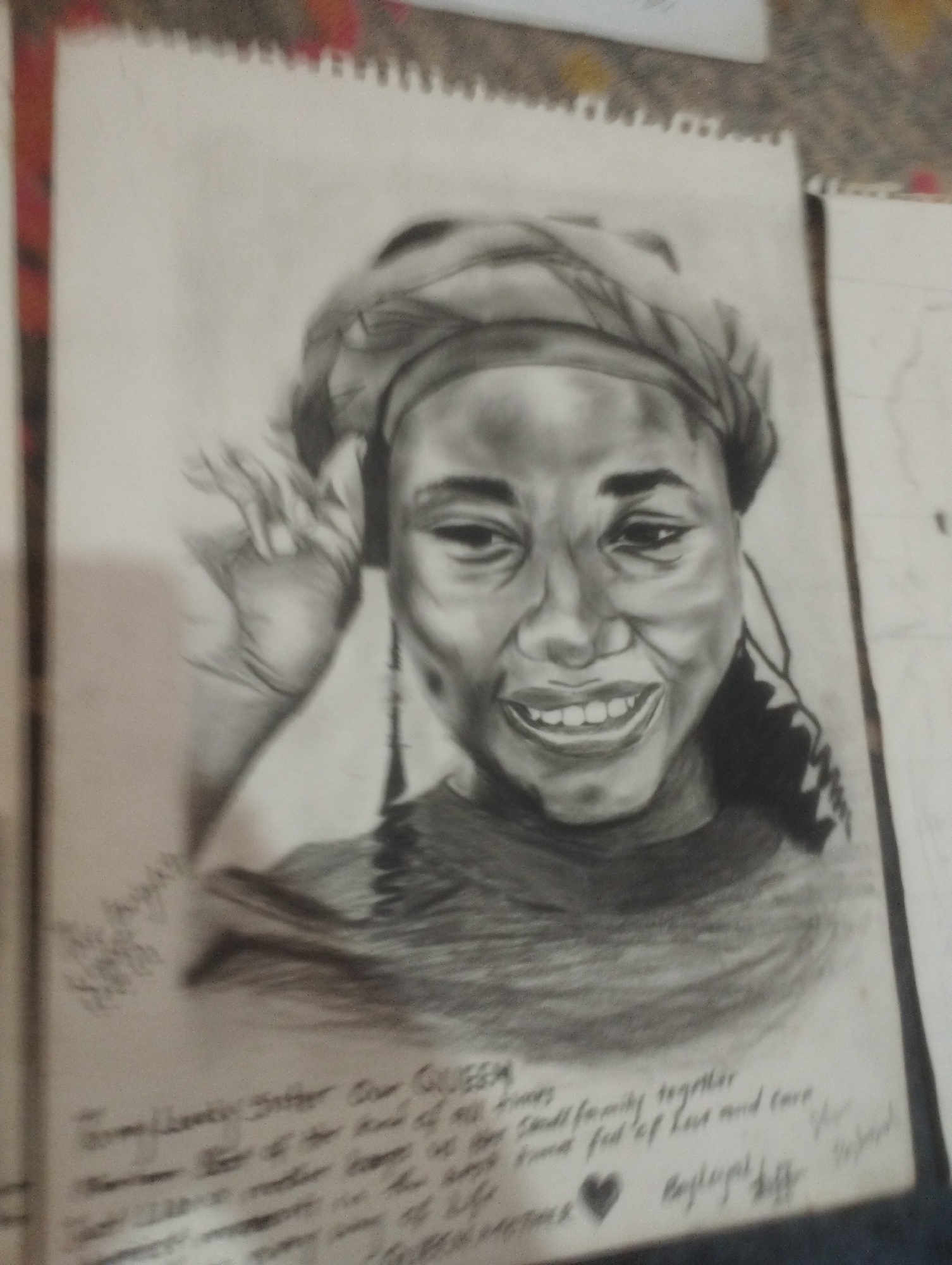 This is a black and white pencil sketch on a sketchbook page depicting a portrait of a young woman. The woman is wearing a headscarf and has a wide smile, showing her teeth. Her left hand is raised near her face. The sketch is detailed, with shading used to create depth and form. There is handwritten text below the portrait, but it is blurry and illegible. The sketchbook page is positioned on a surface with a textured, multi-colored edge visible on the left. The background is out of focus. The image is taken indoors, and there is another sketchbook visible to the right. The overall lighting is soft.