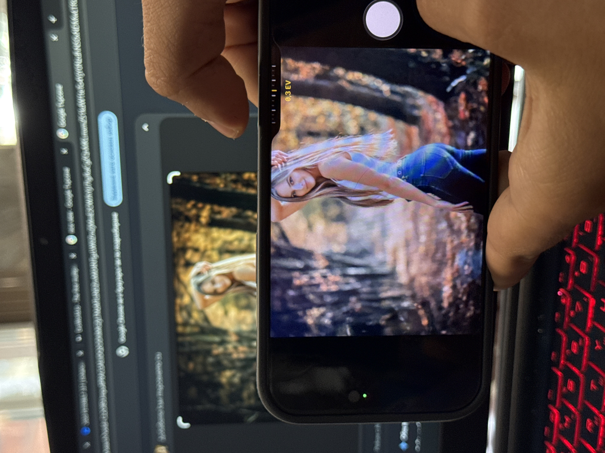 The image captures a person's hand holding a black smartphone, with its screen displaying a photograph of a smiling blonde woman. The woman is lying horizontally on her stomach, looking towards the viewer, with her hair fanned out and wearing a light top and jeans. The background of her photo features warm, blurred autumnal foliage. The smartphone's camera interface is active, showing a white circular shutter button at the top and an exposure compensation slider indicating "0,3 EV" on the left edge. A small green light is visible at the bottom of the phone screen.
In the background, a laptop screen is partially visible on the left, displaying what appears to be Google Chrome with search results. The same image of the blonde woman can be seen partially on the laptop screen, possibly as a wallpaper or a search result. Faint text "Google Chrome" is visible multiple times on the laptop screen. To the right, a section of a red backlit keyboard is visible. The scene is set indoors, likely at a desk, in Varna, Bulgaria.