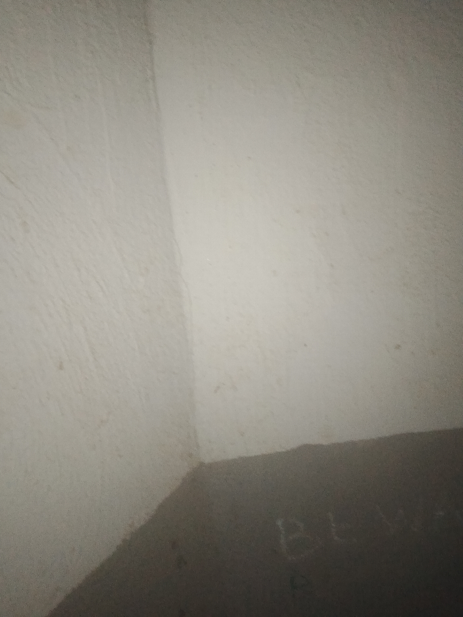 The image shows a dimly lit interior corner of a room with textured, off-white walls. A diagonal shadow falls across the lower portion of the scene, obscuring a portion of the wall. Faint markings, possibly text or graffiti, are visible within the shadowed area, appearing as light-colored scrawls against the dark surface. The overall impression is of an abandoned or neglected space, potentially in a residential or utility building in Jalingo, Nigeria. The lighting suggests the photograph was taken with a flash or a low-light setting, contributing to the grainy quality of the image.