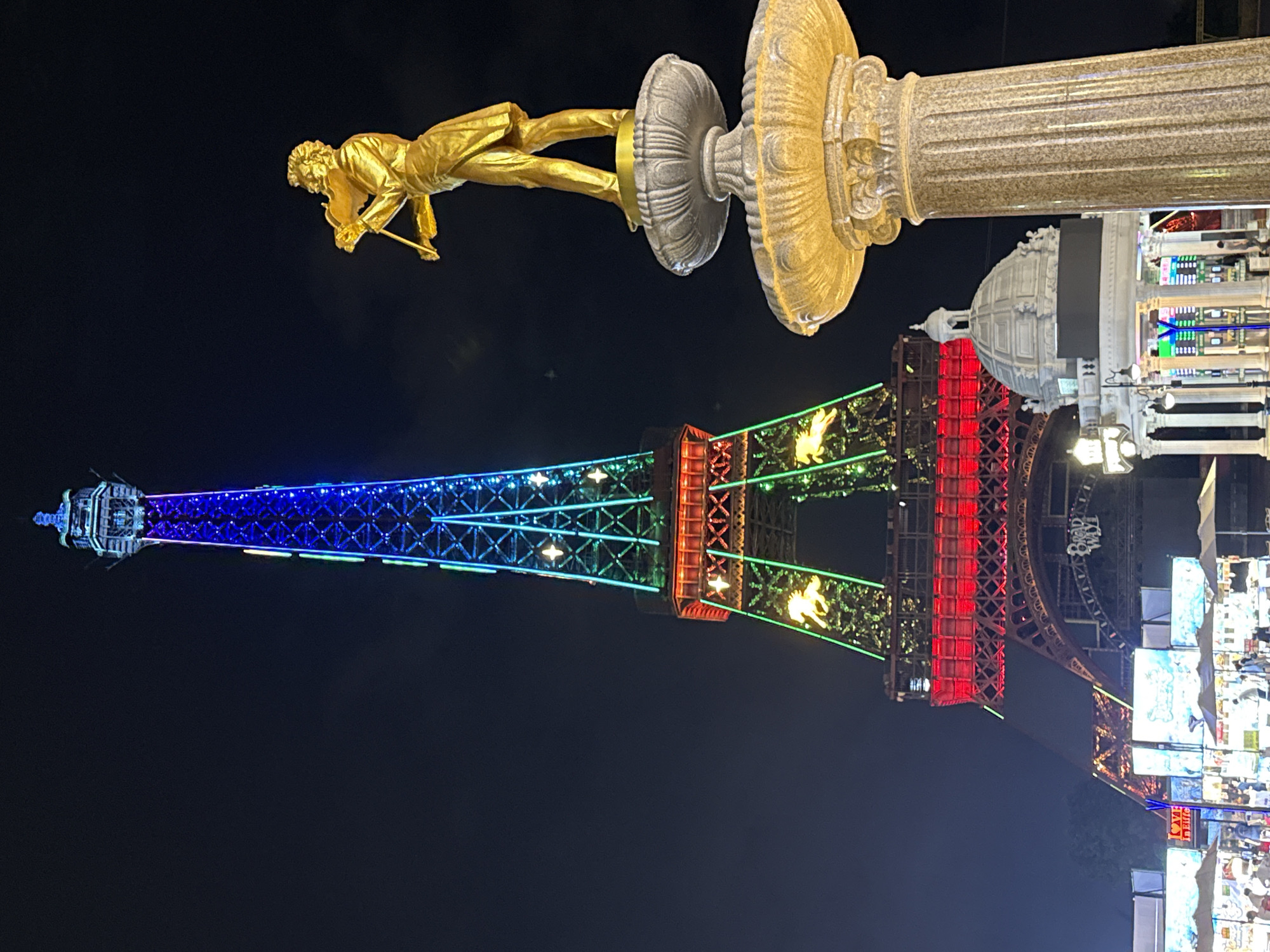This image captures a nighttime view of a replica Eiffel Tower illuminated with colorful lights in the city of Nanshan District, China. A golden statue of a man playing a violin stands atop a fountain in the foreground, to the right of the tower. The tower itself is adorned with red, green, and blue lights, creating a festive atmosphere. Smaller structures and market stalls are visible at the base of the tower, with people browsing the offerings. The overall scene suggests a popular tourist attraction or entertainment venue.