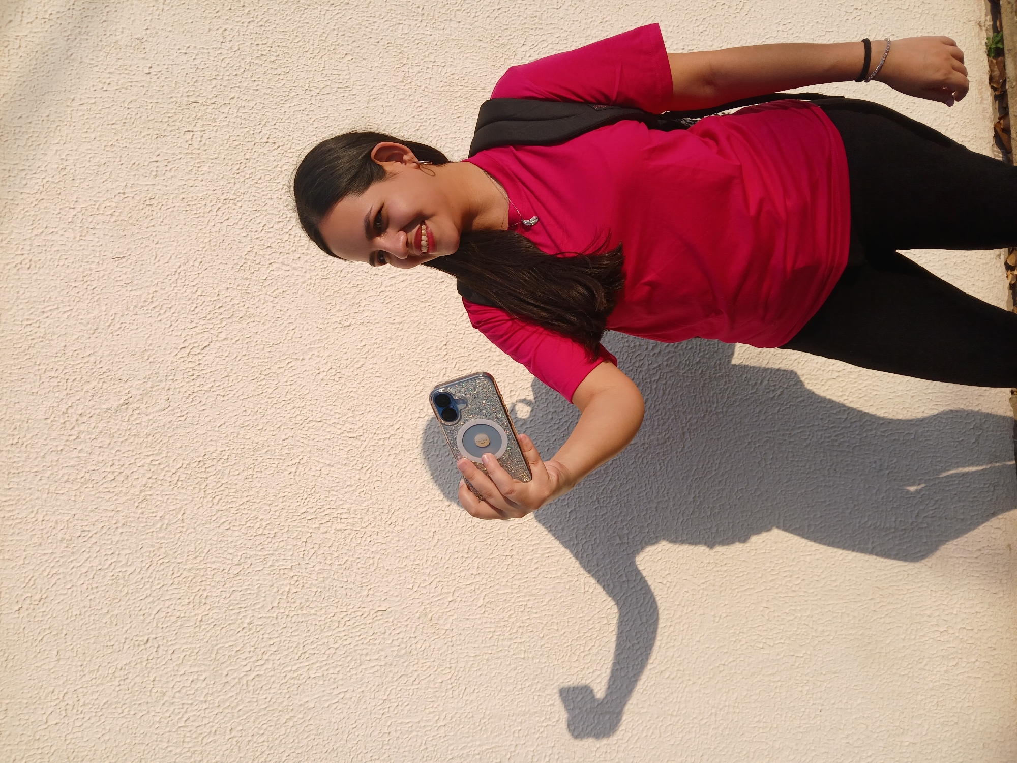 A young woman is holding a smartphone, possibly taking a selfie, in Guayana City, Venezuela. She has long dark hair and is wearing a pink t-shirt and black leggings. A black backpack is visible on her back. She is standing in front of a light-colored textured wall. A shadow of her arm and the smartphone is cast on the wall. The setting appears to be outdoors in daylight.