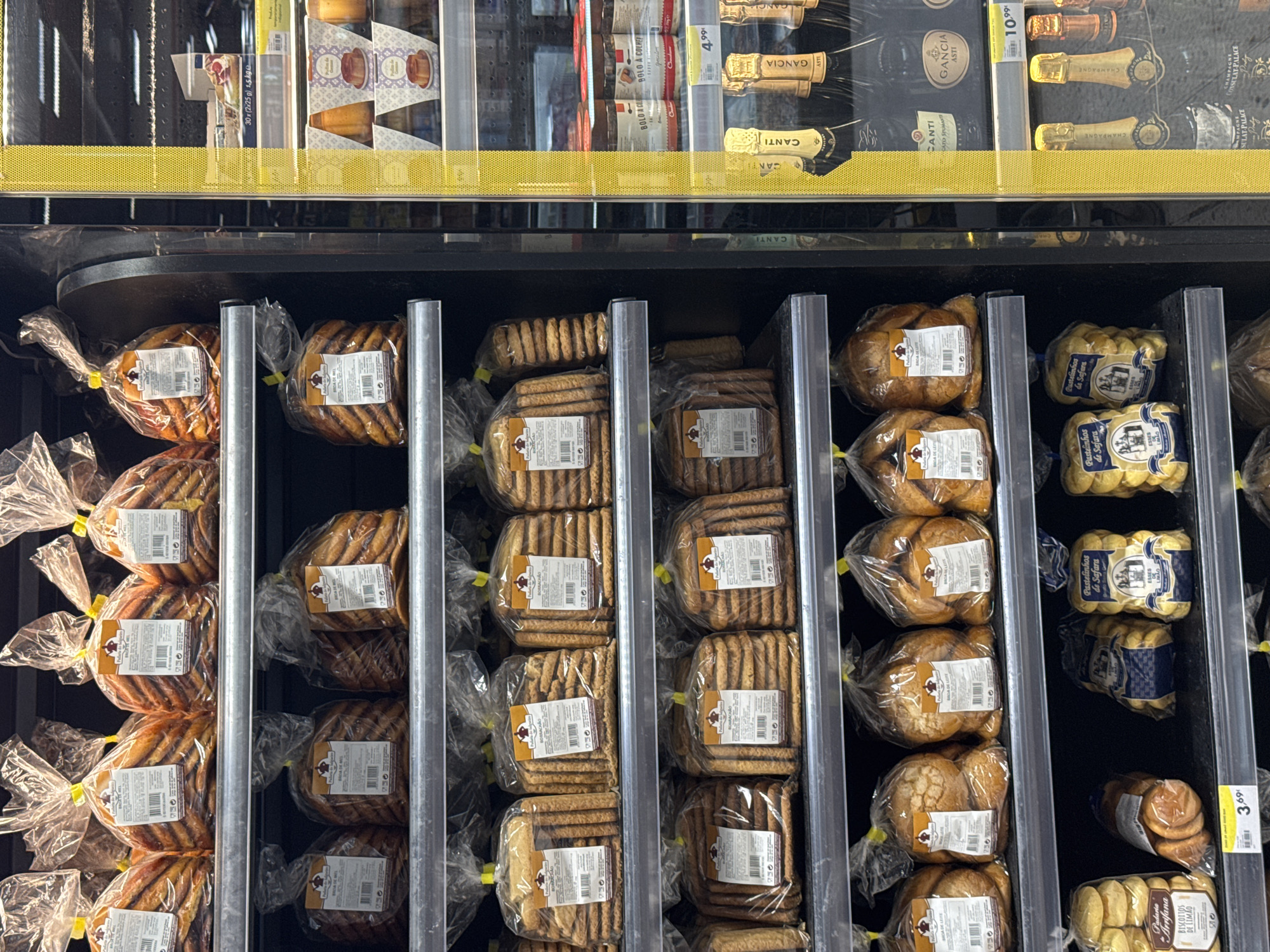 The image shows a refrigerated display case in a supermarket, likely in Castelo Branco, Portugal, based on the presence of Portuguese language packaging. The display is filled with various packaged baked goods, primarily biscuits and cakes, arranged on multiple shelves. The products are predominantly in clear plastic packaging tied with yellow ribbons, with labels displaying brand names, product information, and nutritional details in Portuguese.
On the top shelves, there are large bags of what appear to be biscuits, some with a layered, slightly curved appearance. The labels on these bags show the text "Pão de Ló" and "BRISA DE MEL." Further down, another set of shelves contains bags of biscuits labeled "BORRACHÃO" and also "Pão de Ló."
The lower shelves display different types of baked goods. One shelf has bags of round, bread-like items labeled "BRIGA DE LEITE." Another shelf shows smaller packages of what seem to be shortbread or cookies, labeled "Pao da Avó" and "BISCOITOS DE LIMÃO." There are also bags of a more elaborately shaped pastry labeled "Pastelinhos de Safara."
To the right of the baked goods display, a portion of a yellow, textured panel is visible, which is part of the supermarket's interior design. Beyond this panel, another refrigerated section is visible, containing bottles of sparkling wine, including brands like "CANTY" and "GANCIA ASTI," as well as jars of preserves labeled "BOLO A COLHER." Price tags are also visible, such as "4.99€" and "10.99€."
The overall environment appears to be a clean, well-lit commercial setting. No people are visible in the frame. The focus is entirely on the merchandise within the refrigerated display.