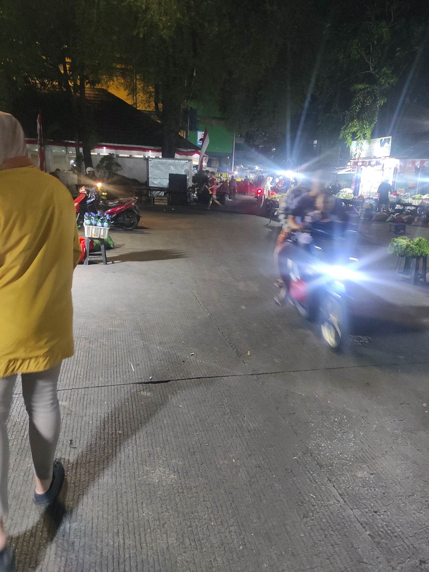 An evening outdoor scene in South Jakarta, Indonesia, depicts a busy street market at night. The foreground features the lower half of a person wearing a yellow jacket and light-colored pants, casting a shadow on the concrete pavement. To the left, a red motorcycle is parked, with a basket of bottled drinks on a small table next to it. In the mid-ground, a motorcycle with bright headlights is in motion, its rider blurred by the speed. Behind this, several market stalls are visible, illuminated by lights, with people browsing and vendors attending to customers. Lush green trees form a canopy over parts of the scene, and there are glimpses of buildings and Indonesian flags in the background. The overall atmosphere is one of a lively nocturnal marketplace.