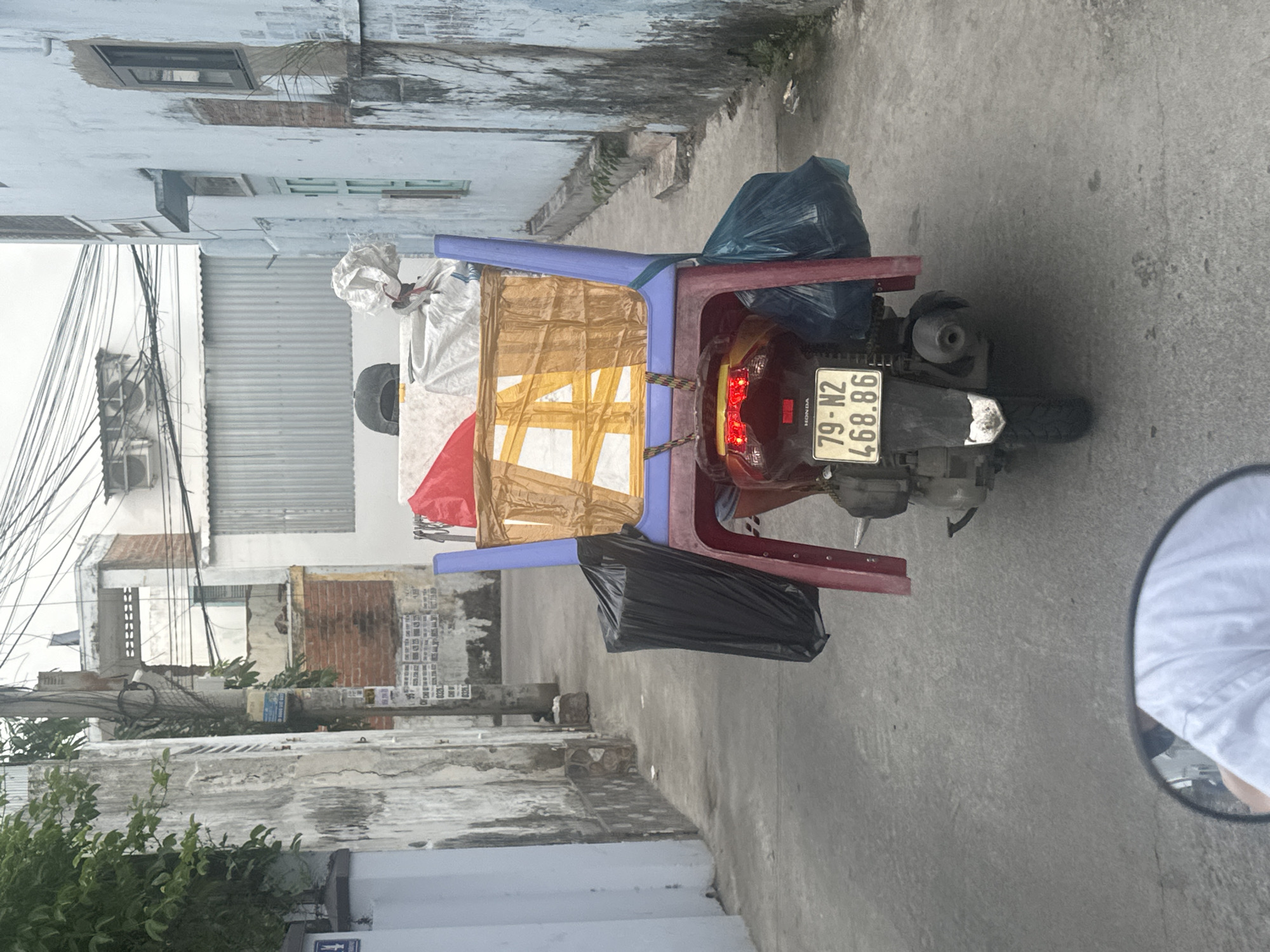The image depicts a motorbike heavily laden with goods traveling down a narrow alleyway in the city of Bac Nha Trang Ward, Vietnam. The motorbike has a license plate that reads "79-N2 468.86." It is carrying an assortment of items, including what appears to be wrapped packages secured with tape and rope, as well as black and blue plastic bags. The items are being transported using a makeshift rack constructed from blue and red plastic chairs. The alley is lined with buildings of varied construction, including a mix of brick, concrete, and corrugated metal. Power lines are visible overhead, adding to the urban landscape. The overall scene suggests a bustling, informal economy. A person is visible in the motorbike's rearview mirror.