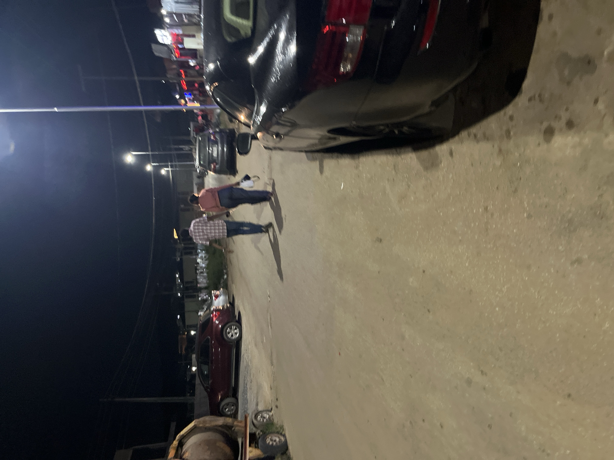 At night in Abuja, Nigeria, two individuals, a man and a woman, are walking away from the camera down a dirt road. The man, wearing a plaid shirt and blue jeans, walks slightly ahead of the woman, who is dressed in a salmon-colored top and dark trousers and carries a white bag. They are walking past several parked vehicles, including a dark-colored sedan and a red SUV. In the foreground, a black car is parked on the right side of the road. The road is unpaved and appears to be a side street with streetlights illuminating the scene. Businesses with illuminated signs are visible in the background on the right side of the road.