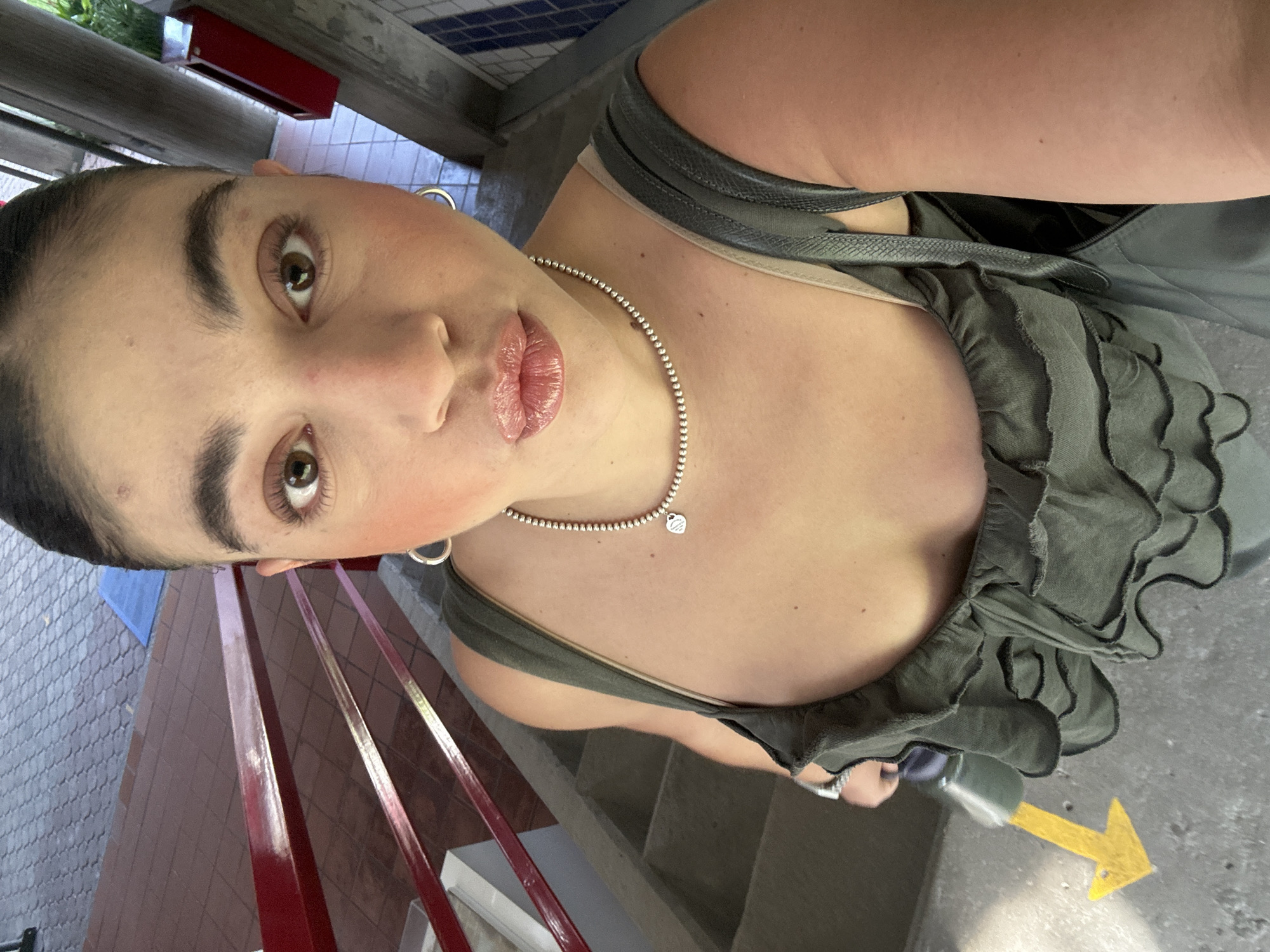 A close-up, angled selfie captures a woman posing in what appears to be an outdoor or semi-outdoor public setting in San Bernardino Tlaxcalancingo, Mexico. The woman is the central subject, looking directly into the camera with her head tilted. Her dark hair is pulled back, revealing her prominent eyebrows and brown eyes. She wears light makeup, including blush and glossy, slightly pouting lips.
She is dressed in an olive green top with delicate ruffled detailing across the chest, and the strap of a thin-strapped undergarment is visible beneath. Around her neck, she wears a silver beaded necklace featuring a small, reflective charm, possibly branded, though the text is illegible. Small silver hoop earrings adorn her ears.
The background elements suggest a utilitarian or public space. To her left, red-painted railings or bars run diagonally upwards. Behind her, a wall features light-colored tiles, possibly blue and white or similar hues. The ground is concrete, and notably, a yellow arrow pointing downwards is painted on it in the lower right corner of the frame. The bright, clear lighting indicates that the photo was taken during the daytime. The overall impression is a casual, posed portrait.