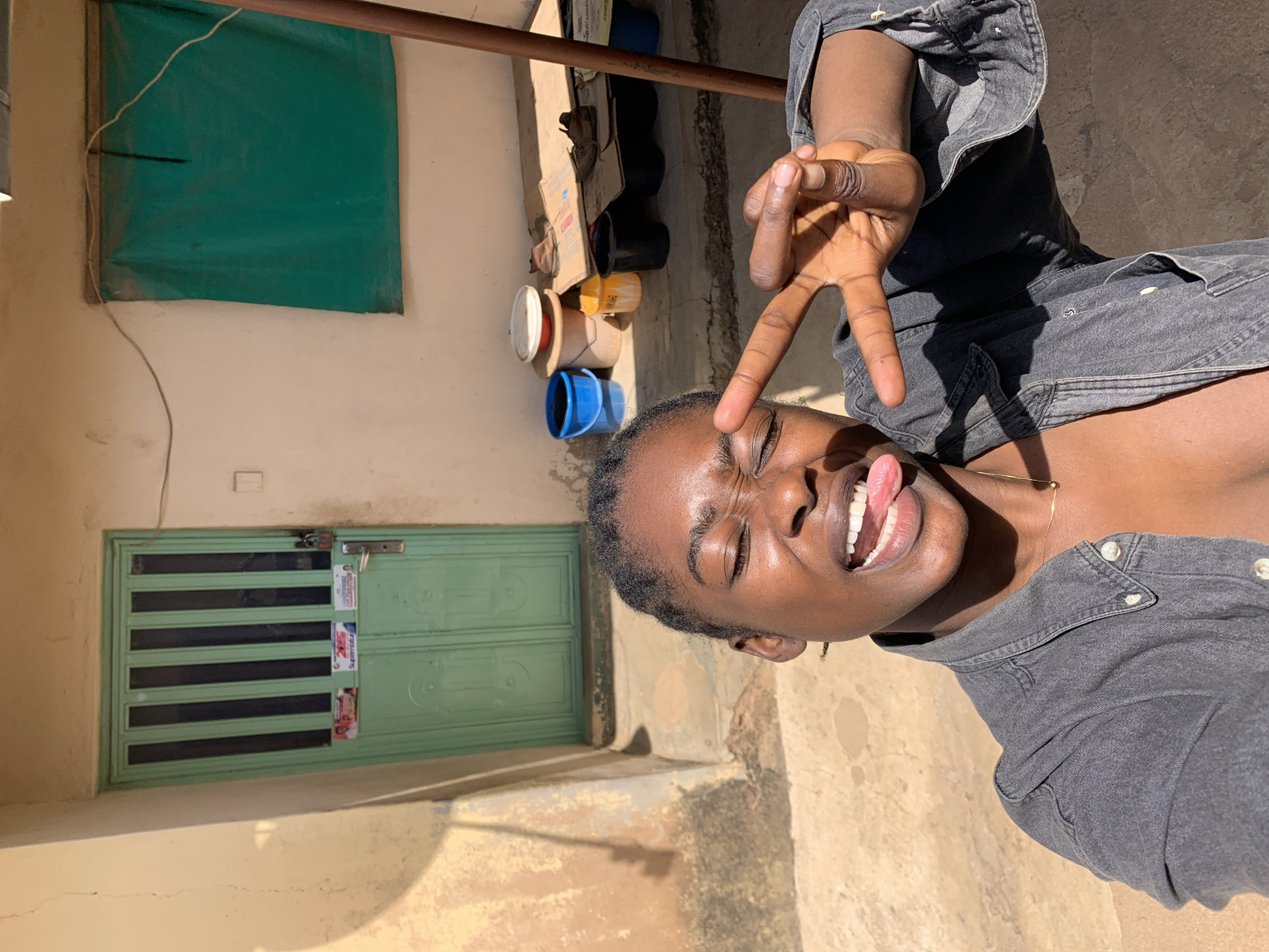 A person stands outside a building in Rafin Par, Nigeria. The building has a light green door and a covered window. Various objects, including containers and shoes, are on the ground near the building. The person is holding up a peace sign and sticking out their tongue. The ground appears to be made of concrete or stone.