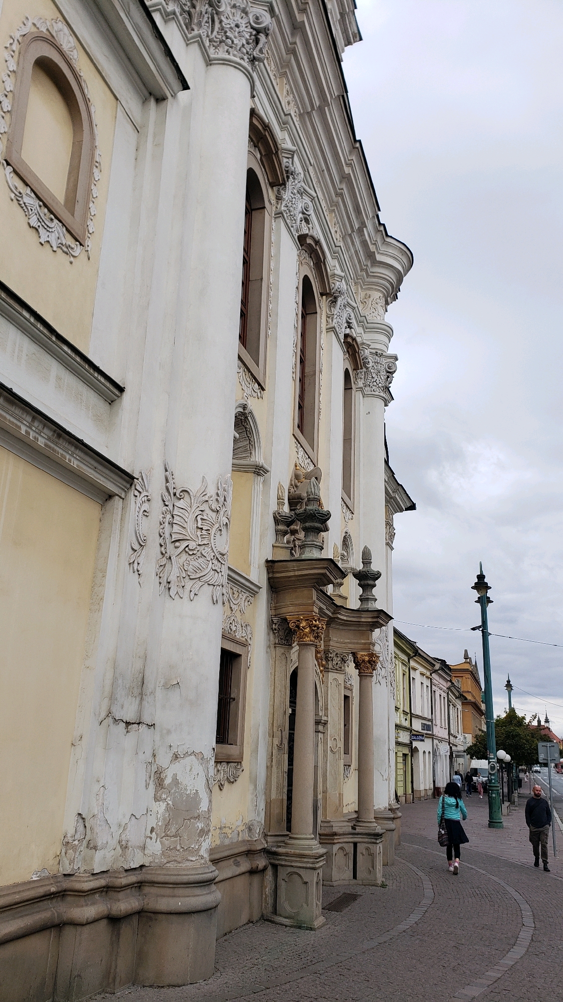 The image shows a street scene in Prešov, Slovakia, featuring the facade of a grand, ornate building on the left and a street with several people walking on the right. The building on the left is light yellow with extensive white decorative plasterwork, including columns, arches, and sculpted elements. The plaster on the lower section of the building shows signs of wear and cracking. The building's architecture appears to be of historical significance, possibly a church or a significant public building.
The street is paved with cobblestones in a curved pattern along the building's facade. The right side of the image shows a sidewalk with several people. In the foreground, a woman in a teal jacket and a dark skirt walks away from the camera. Further down the street, a man in a dark jacket and pants walks towards the camera. Other pedestrians are visible in the distance. The street is lined with a row of colorful buildings with shops or residences. A lamppost with multiple lights stands on the sidewalk. The sky is overcast, suggesting a cloudy day. The overall impression is of a historic urban environment with everyday activity.