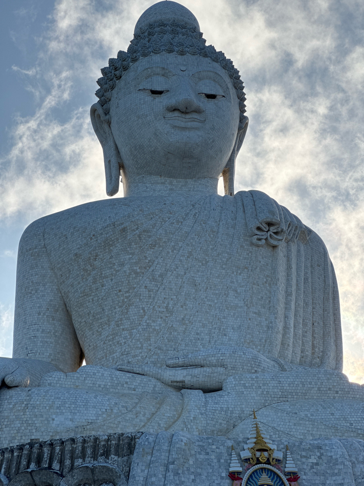 The image captures a low-angle, full-body view of the Phuket Big Buddha statue, a prominent landmark in Karon, Thailand. The statue, constructed from white marble, is meticulously tiled, giving it a textured appearance. It is depicted in a seated meditative pose, with hands resting in its lap. The statue's face, serene and impassive, is turned slightly towards the viewer. Behind the statue, the sky is visible, characterized by a mix of bright sunlight and scattered, wispy clouds. The lower section of the image shows the base of the statue, which is adorned with decorative architectural elements, including a tiered, golden spire. The overall scene is bathed in natural light, highlighting the grandeur of the statue against the backdrop of the sky.