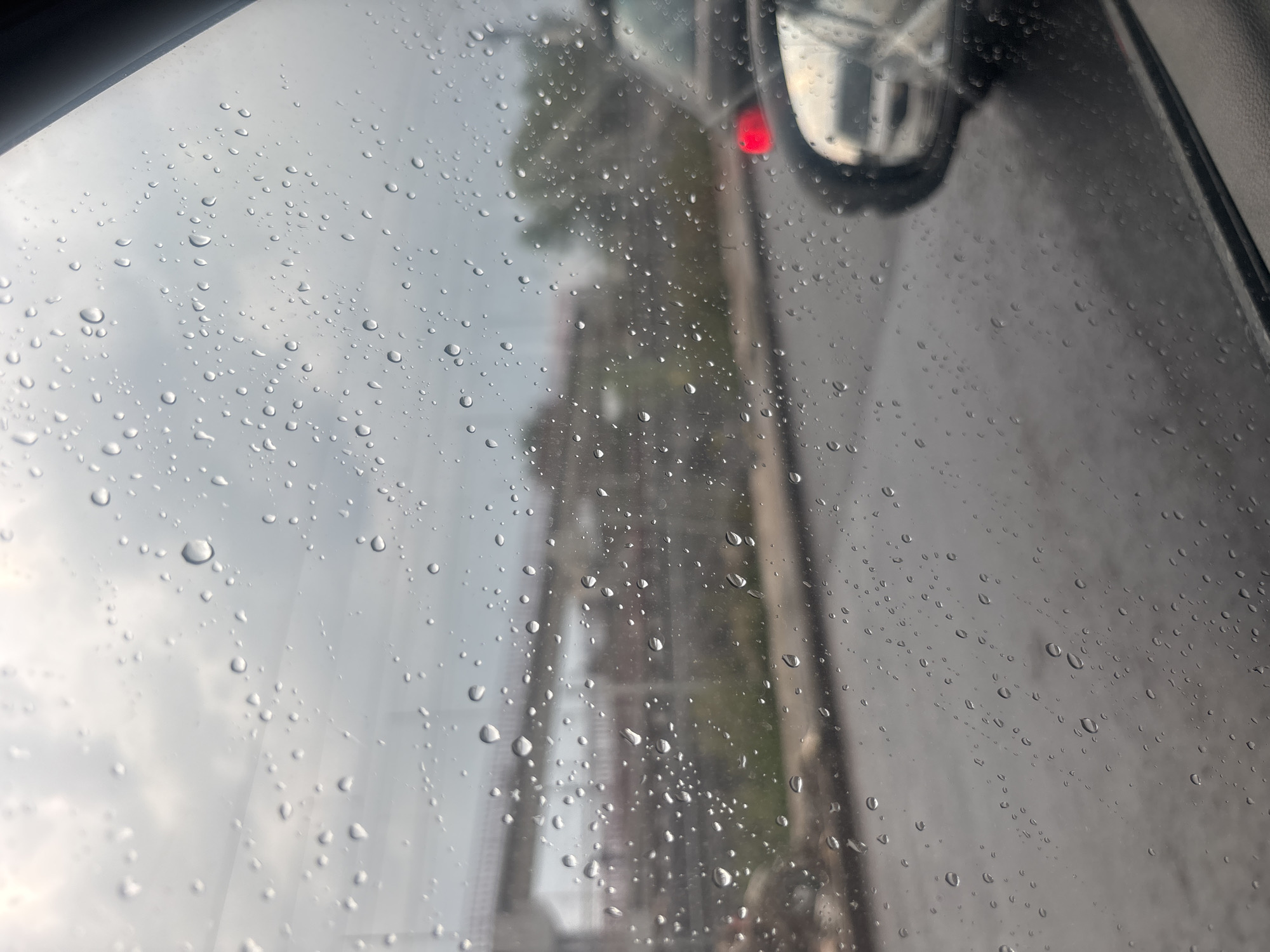 The image shows a view through a rain-covered car window. The raindrops obscure the scene outside, but a red light is visible in the reflection of a car side mirror. In the background, a blurred building or structure is visible, along with power lines. The overall impression is of a rainy day in an urban setting. There are no people visible. The city of Tultitlán, Mexico cannot be determined from this image.