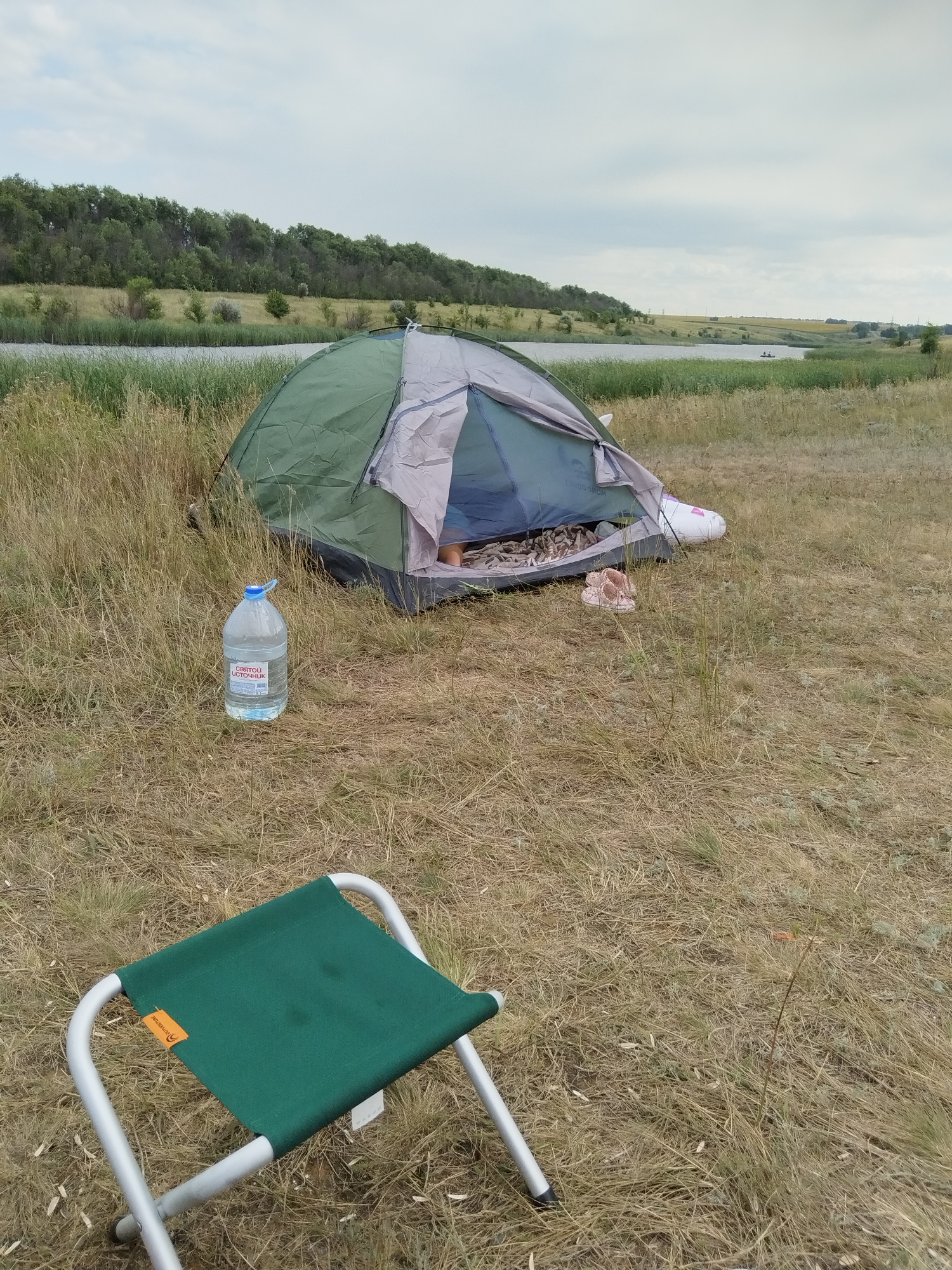 In Parfenovka, Russia, a green and grey tent is set up on dry, grassy terrain near a body of water. A large bottle of water with a red label is placed on the ground in front of the tent. A green folding stool is positioned in the foreground of the image. The landscape features a tree line bordering the water and a cloudy sky overhead.