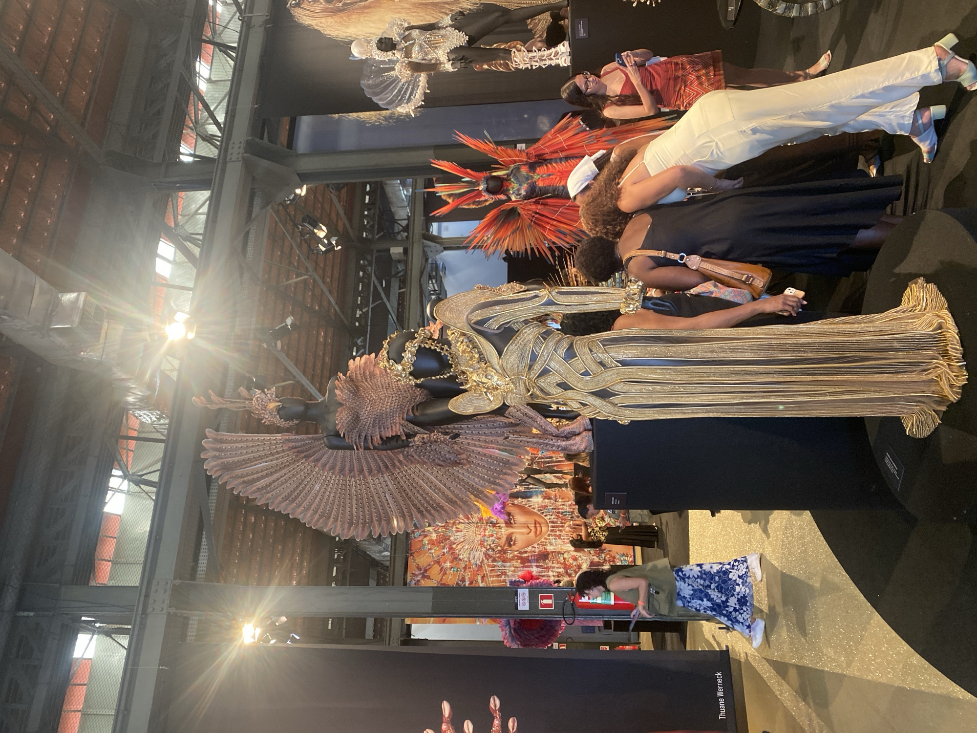 The image shows an indoor display with several mannequins dressed in elaborate costumes. The architecture of the building is industrial, with exposed metal beams and a high ceiling. There are multiple people in the scene, including some observing the mannequins and others taking photos. A large colorful image is displayed in the background. The lower portion of the photo displays the name "Thuane Werneck" on a sign. Based on the costumes, display, and name, the location is likely connected to the Rio Carnival in the city of Rio de Janeiro, Brazil.