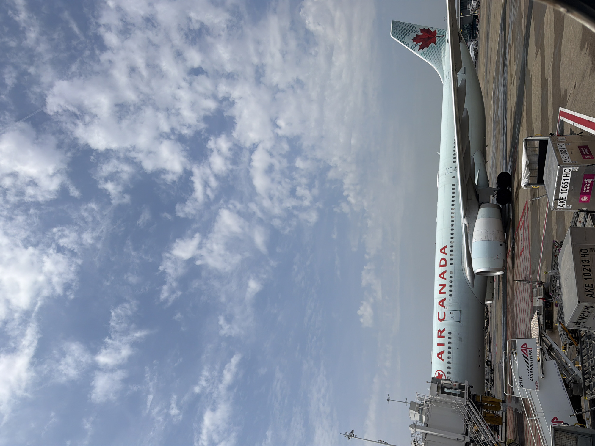 The image captures an Air Canada aircraft parked at an airport gate, presumably for boarding or deplaning. The plane is positioned parallel to the terminal, with the "AIR CANADA" logo prominently displayed along its fuselage. Loading equipment and ground personnel are visible near the aircraft, indicating ongoing ground operations. The sky above is partly cloudy.
The text "Zaventem, Belgium" does not appear in the image and is not inferrable from the other objects in the image.