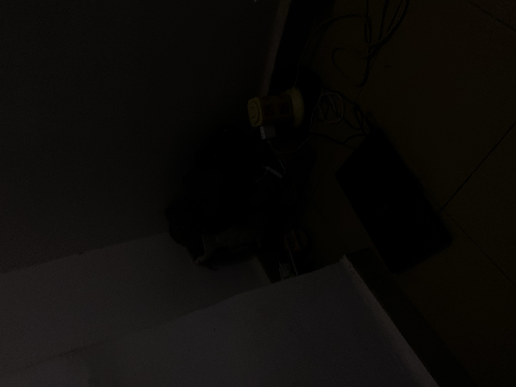 In a dimly lit room, likely a bedroom or storage area, a dark rectangular object, possibly a tablet or a flat-screen television, rests on a wooden floor. To the right, a yellow cylindrical object, resembling a small fan or a humidifier, is plugged into a white power adapter. Various black and white cables are scattered around the power adapter and the yellow object. In the background, a dark bag is slumped against the corner of the room, where two walls meet. The lighting is very low, casting deep shadows and obscuring finer details. The overall impression is one of a cluttered, utilitarian space. The location of Howayi, Nigeria, is not visually discernible from the provided image.