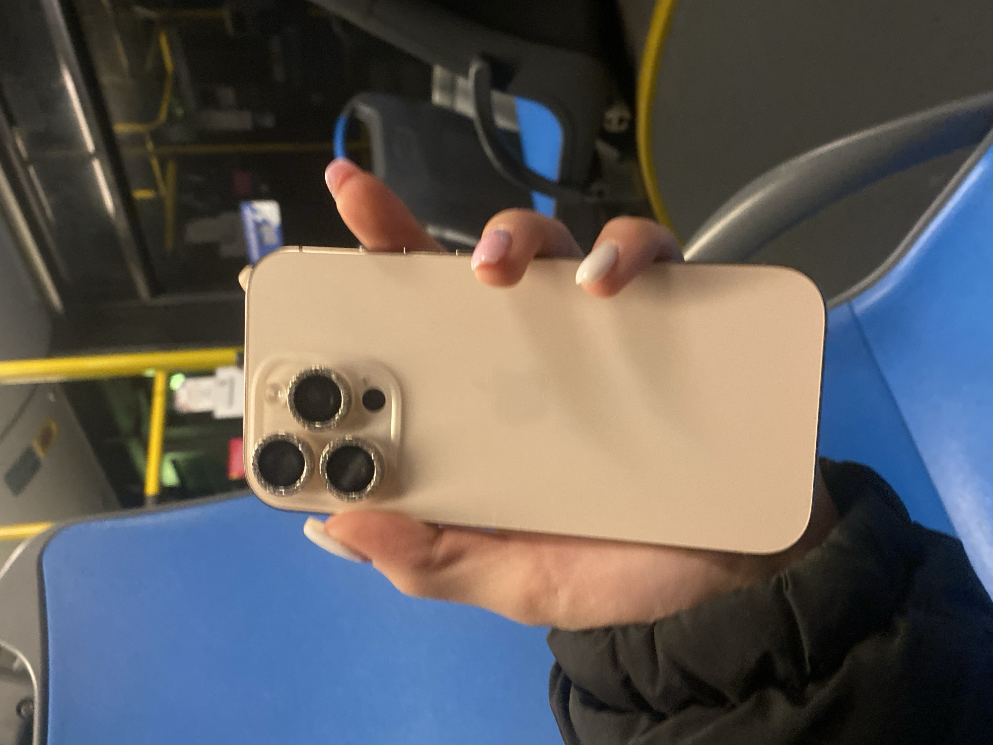 A hand holds a gold-colored iPhone with a triple-lens camera system. The phone appears to be an iPhone 13 Pro or later model. The background shows the interior of a bus, identifiable by the blue plastic seating, metal support bars, and overhead storage racks. The location appears to be a bus in Zvezditsa, Bulgaria.