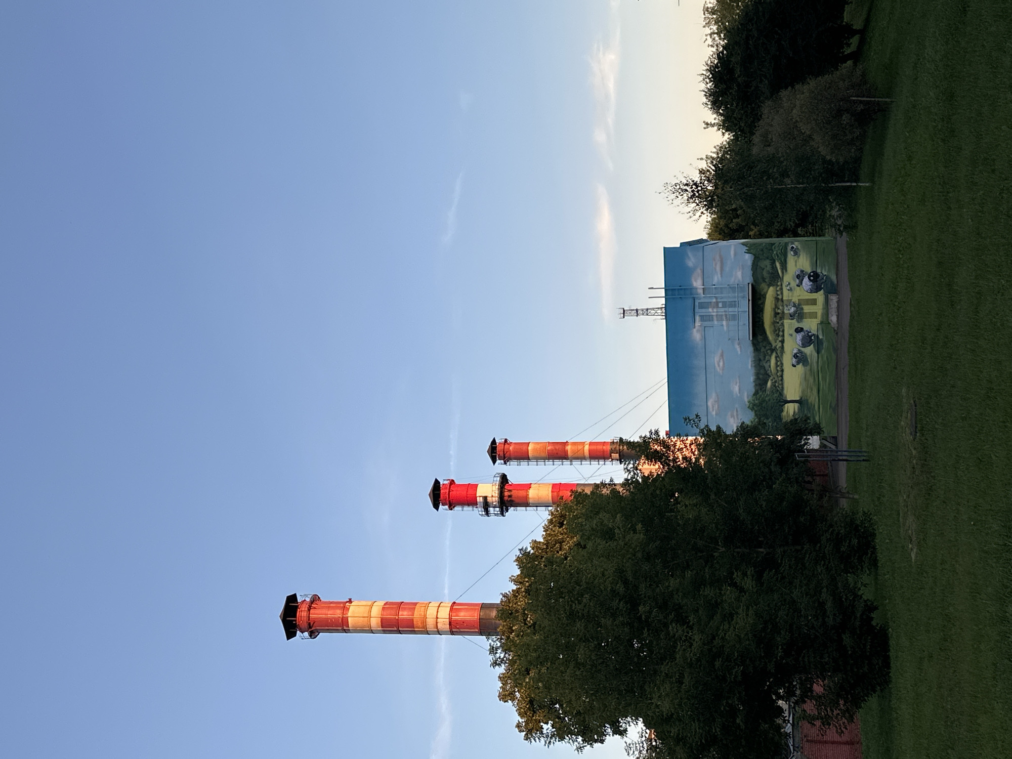 The image shows a view of three industrial chimneys in the city of Minsk, Belarus. The chimneys are tall and cylindrical, with red and white stripes. Two of the chimneys are closer to the foreground and are taller than the third one, which is further in the background. In front of the chimneys, there is a building with a mural painted on its side. The mural depicts a rural landscape with sheep grazing in a field under a blue sky with white clouds. A large green tree is visible in the foreground, obscuring a portion of the chimneys. The area around the building and chimneys is covered in green grass, indicating a park or open area. The sky is clear with a gradient of blue, transitioning to lighter shades near the horizon.