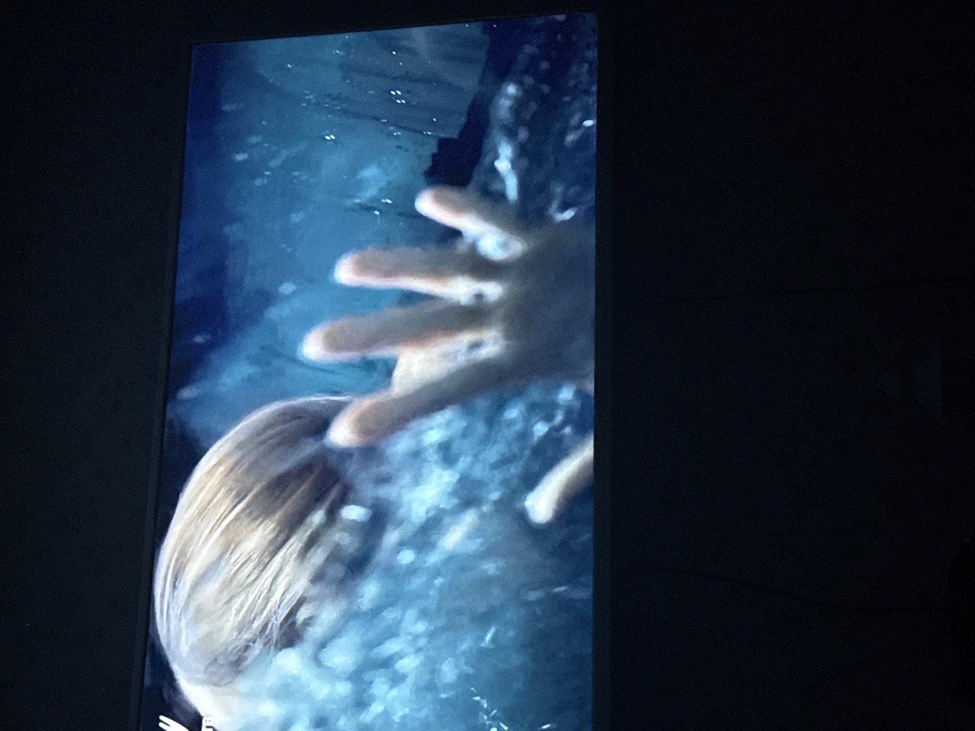 The image shows a scene from what appears to be a television or monitor. The scene appears to depict a person, possibly female, with blonde hair underwater. A hand is reaching towards the viewer, surrounded by bubbles and water. The scene is predominantly blue and white, suggesting underwater conditions. The location of the image is not discernible from the provided visual data.