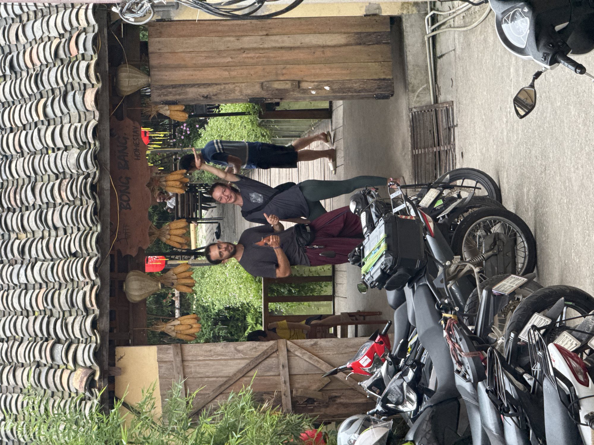 The image shows a group of three people posing in front of a building with a tiled roof and a sign that reads "Bong Bang Homestay." The location appears to be in Yên Minh Commune, Vietnam. A man is standing on the left, a woman is striking a playful pose in the middle, and another man is leaping in the background, all seemingly interacting. Several motorcycles are parked on the right side of the frame, next to the people. Vegetation is visible in the foreground and background.