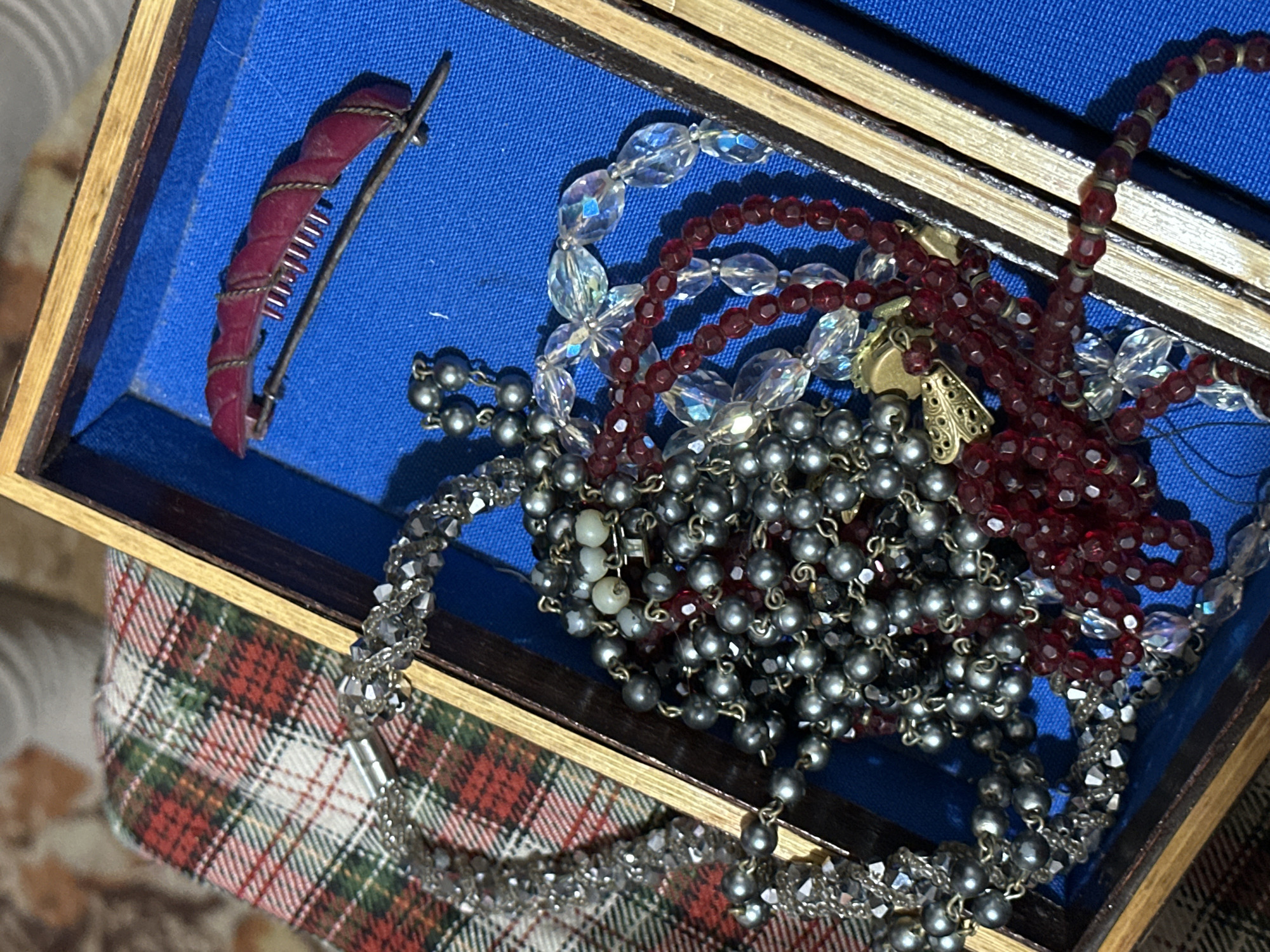 A wooden jewelry box, lined with blue fabric, sits on a surface covered with a plaid cloth. Inside the box are various pieces of jewelry, including red beaded necklaces, clear crystal bead necklaces, and a grey pearl necklace. A red and metal brooch is positioned in the upper left corner. The location is not identifiable from the image.