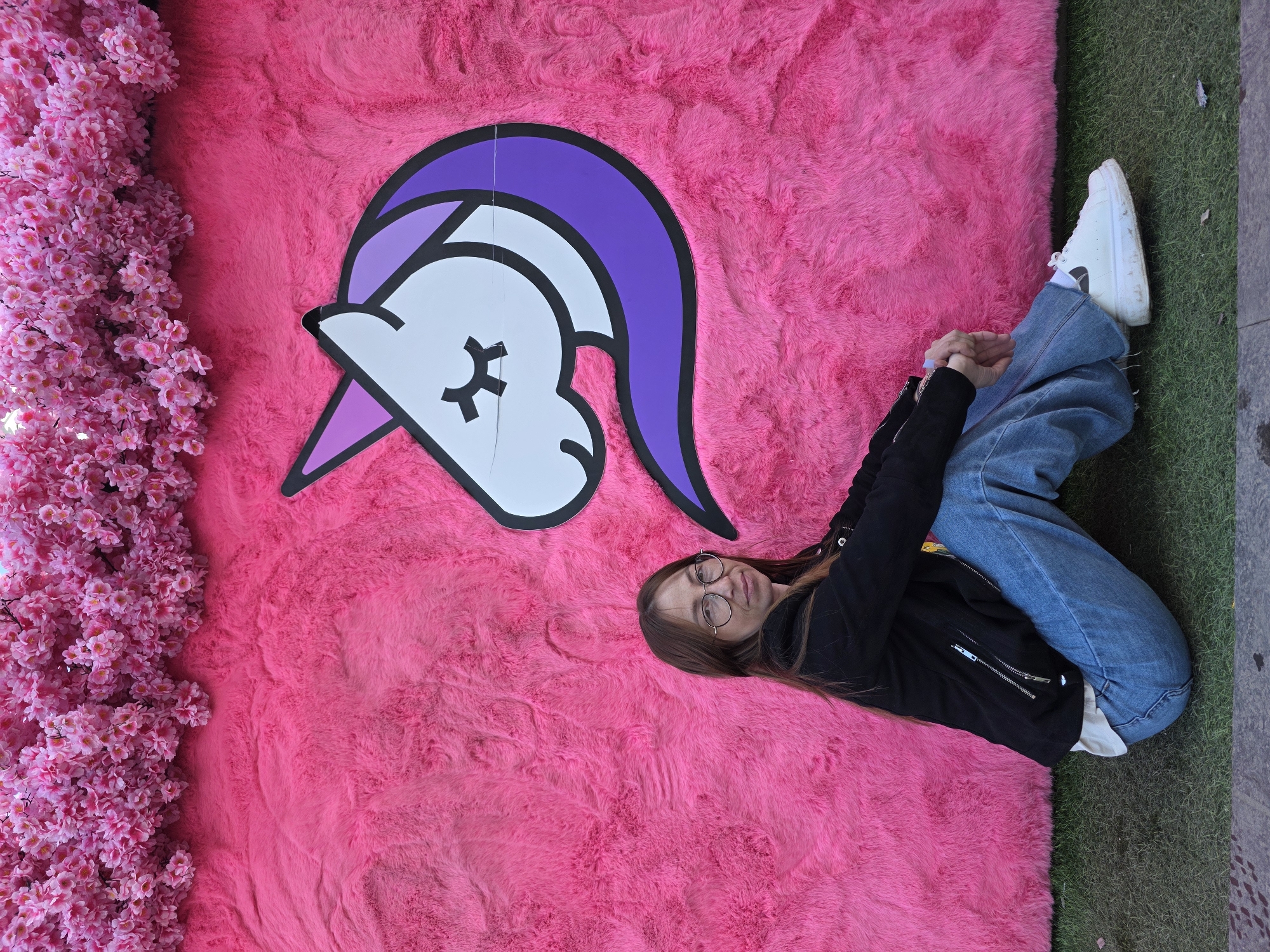 An individual is seated on artificial green grass in front of a promotional backdrop. The backdrop features a wall covered in bright pink faux fur, topped with a dense arrangement of artificial pink cherry blossoms. A large graphic of a white unicorn head with a purple mane and horn, featuring closed eyes, is mounted on the pink fur to the right of the person.
The individual, appearing female, has long light brown hair and wears round eyeglasses. They are dressed in a black zip-up jacket over a light-colored shirt, loose-fitting blue jeans, and chunky white sneakers. Their legs are bent and crossed, with hands clasped in front. The individual is looking directly at the camera with a neutral expression. The very bottom of the image shows a strip of dark concrete or asphalt bordering the artificial grass. The specific location within Votkinsk, Russia, cannot be determined from the visual elements of this artificial setup.