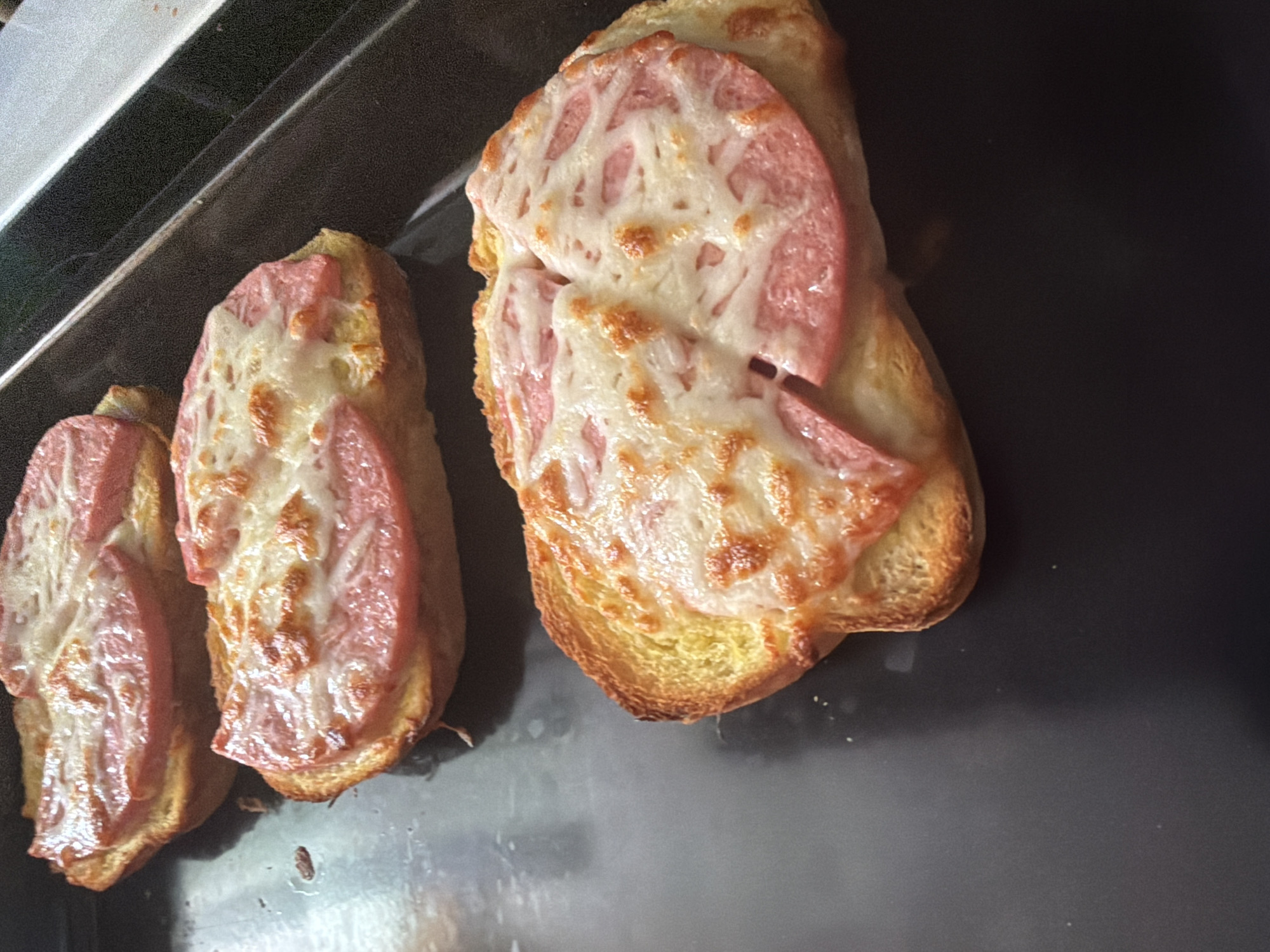 The image displays three individual pieces of toasted bread, each topped with a slice of pink meat, resembling ham or sausage, and melted cheese. The cheese exhibits a bubbly, golden-brown texture in places, indicating it has been recently cooked or broiled. The bread underneath is golden-brown and appears crispy. These items are arranged on a dark, reflective surface, likely a baking sheet or oven tray. A metallic, possibly silver, edge of an appliance is visible in the upper left corner of the frame, suggesting an indoor kitchen setting. No people or visible text are present in the image. The scene captures freshly prepared food, possibly a snack or appetizer, in Dabene, Bulgaria.