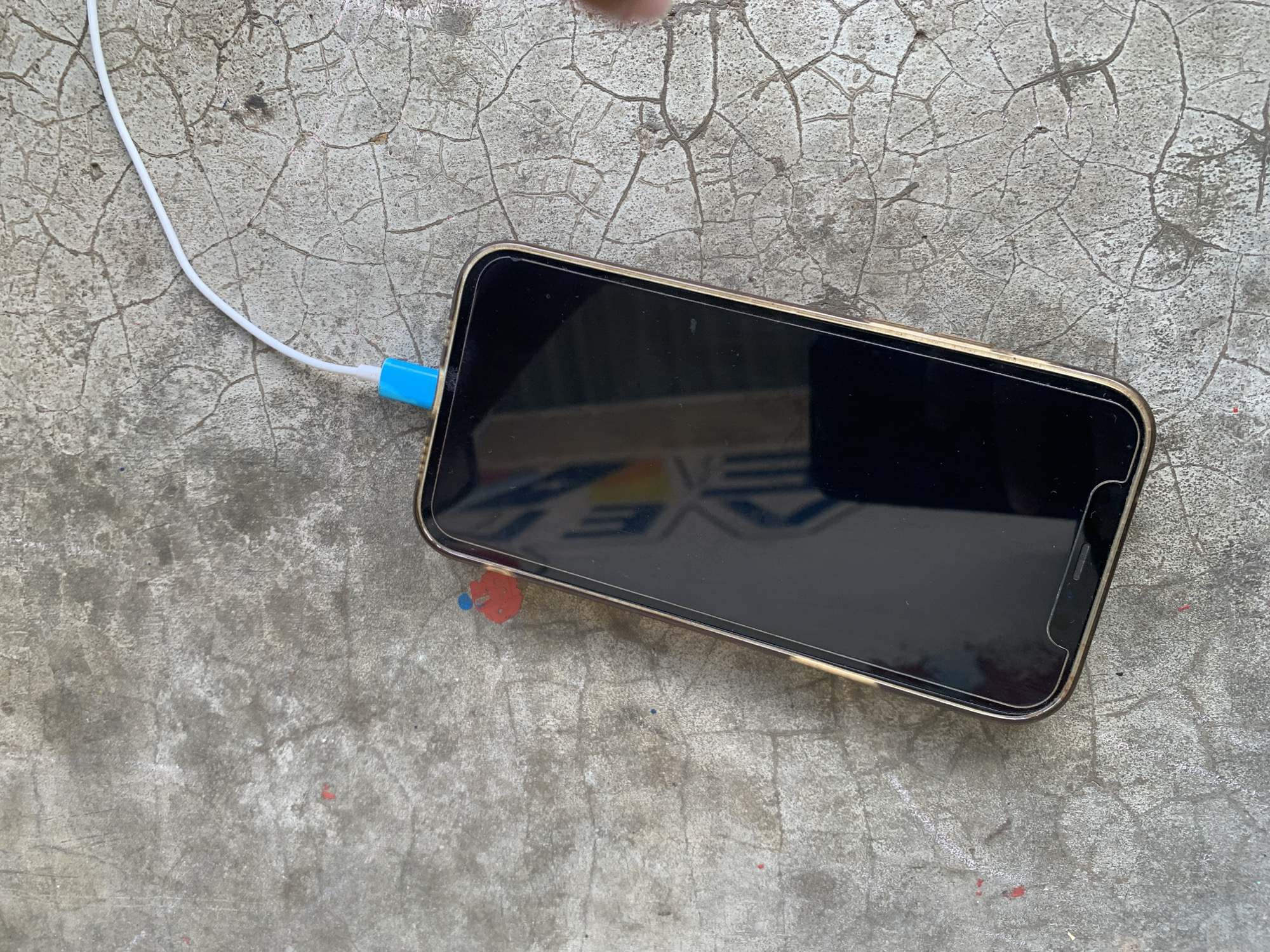 A smartphone is depicted charging on a cracked, gray concrete surface, likely outdoors in Santa Ana, Venezuela. The phone, resembling an iPhone model with a prominent notch and a screen protector, features a golden or bronze-colored metallic frame. A white charging cable, distinguished by its bright blue connector, is plugged into the phone's port.
The phone's screen is dark, showing reflections of its surroundings, including what appears to be part of a mirrored logo or sign with fragmented letters like "AVE" or "EVE." The concrete surface beneath the phone is heavily textured with numerous fine, spiderweb-like cracks, suggesting age or wear. Small, faded specks of red and blue paint or residue are visible on the concrete, particularly in the lower right portion of the frame. Bright, natural lighting illuminates the scene, indicating daytime. A small part of a human finger or thumb can be seen at the very top edge of the image, suggesting a person's presence, perhaps holding or adjusting the phone. The scene captures a moment of everyday technology interacting with a rough, natural setting.