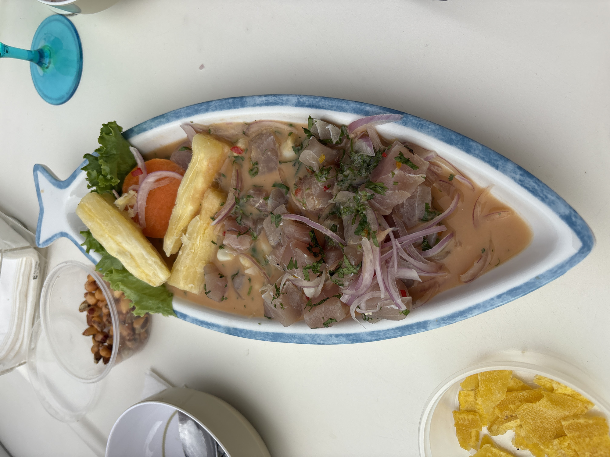 A high-angle, close-up shot captures a Peruvian ceviche dish presented in a white, fish-shaped ceramic boat with blue trim, sitting on a white table. The ceviche features diced raw fish, red onion slivers, chopped herbs, and a creamy orange sauce. Alongside the ceviche are two pieces of cooked cassava and a leaf of lettuce. To the left of the dish, a small clear plastic container holds corn nuts, and another clear container to the lower left contains yellow plantain chips. In the background, a blue wine glass is visible, and a stack of white paper napkins sits behind the ceviche. The scene is set in a restaurant or outdoor dining area in La Punta, Peru.