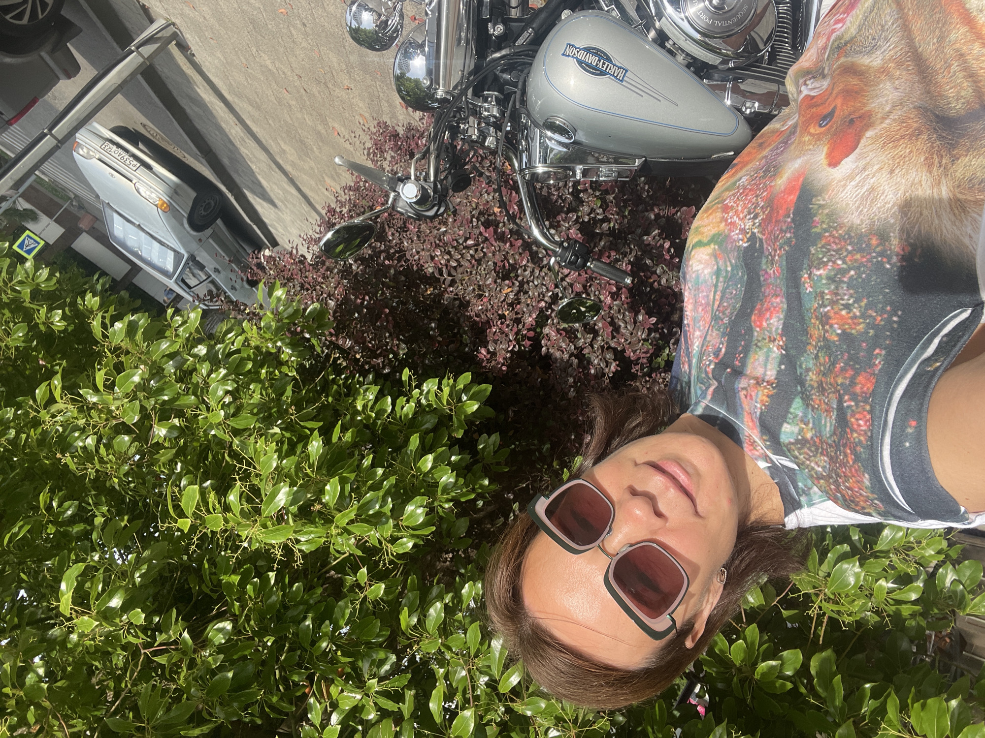 A woman with brown hair and sunglasses is taking a selfie outdoors, with a motorcycle and a car visible in the background. She is wearing a t-shirt with a nature-themed print. The scene appears to be a sunny day. The license plate on the car indicates it is in Russia, and a sign visible in the background suggests a possible location in Sochi.