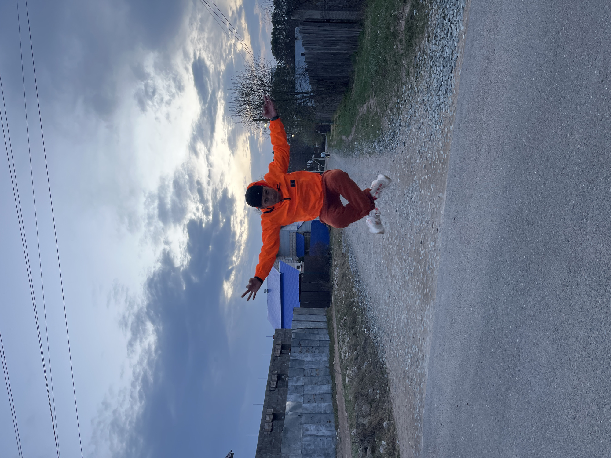 This image, set in Chaikovsky, Russia, captures a dynamic moment of a young man mid-air, performing a jump or "flying" pose. He is positioned centrally, dressed in a bright orange hooded jacket, matching orange pants, and white sneakers, with a dark beanie or cap completing his attire. His arms are spread wide and slightly bent at the elbows, with hands open, and his legs are also outstretched, conveying a sense of energy and exuberance.
The background depicts a semi-urban or rural landscape under a cloudy sky, which shows varying shades of gray and white, suggesting an overcast late afternoon or early evening. Bright patches of sky are visible through the clouds, indicating some light breaking through. Several prominent power lines stretch across the upper left side of the frame. The ground beneath the man is a mix of an unpaved, gravelly path on the left and an asphalt road on the right. Further back, on the right, there's a wooden fence bordering an area with several trees, some of which are bare, indicating either late autumn or early spring. To the left and further back, various structures are visible, including a building with a striking blue roof and another partially built or unfinished structure made of concrete blocks. The overall lighting is soft and diffused. There is no visible text in the image.