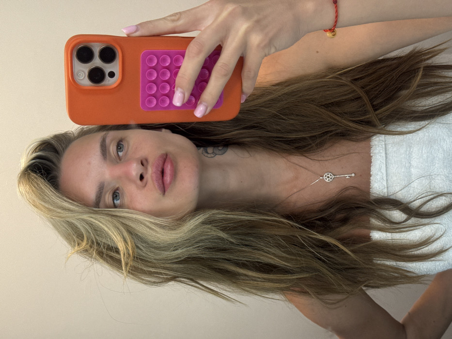 A fair-skinned woman poses for a selfie. She's holding an orange phone case with a pink, suction cup grip. Her long, blonde hair is tousled. She has light eyes, and full lips. A silver necklace with a key pendant rests on her chest. A red string bracelet adorns her wrist. A glimpse of a white towel or robe is visible, suggesting a casual or post-shower setting. She has a visible tattoo on her neck. The image is rotated for a different perspective.