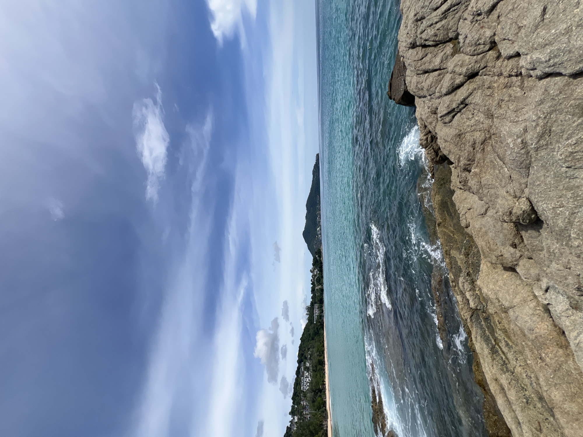 The image depicts a scenic coastal view of Choeng Thale, Thailand. In the foreground, rugged, weathered rocks form a natural barrier against the incoming turquoise water, where small waves crash and create white foam. The middle ground is occupied by the clear, blue-green ocean that stretches towards the horizon. Along the coastline in the distance, a sandy beach is visible, backed by lush green hills dotted with white buildings and trees. A prominent, rounded mountain rises from the sea in the far distance. The sky above is a vibrant blue, adorned with scattered white clouds. The overall impression is a serene and picturesque tropical landscape.