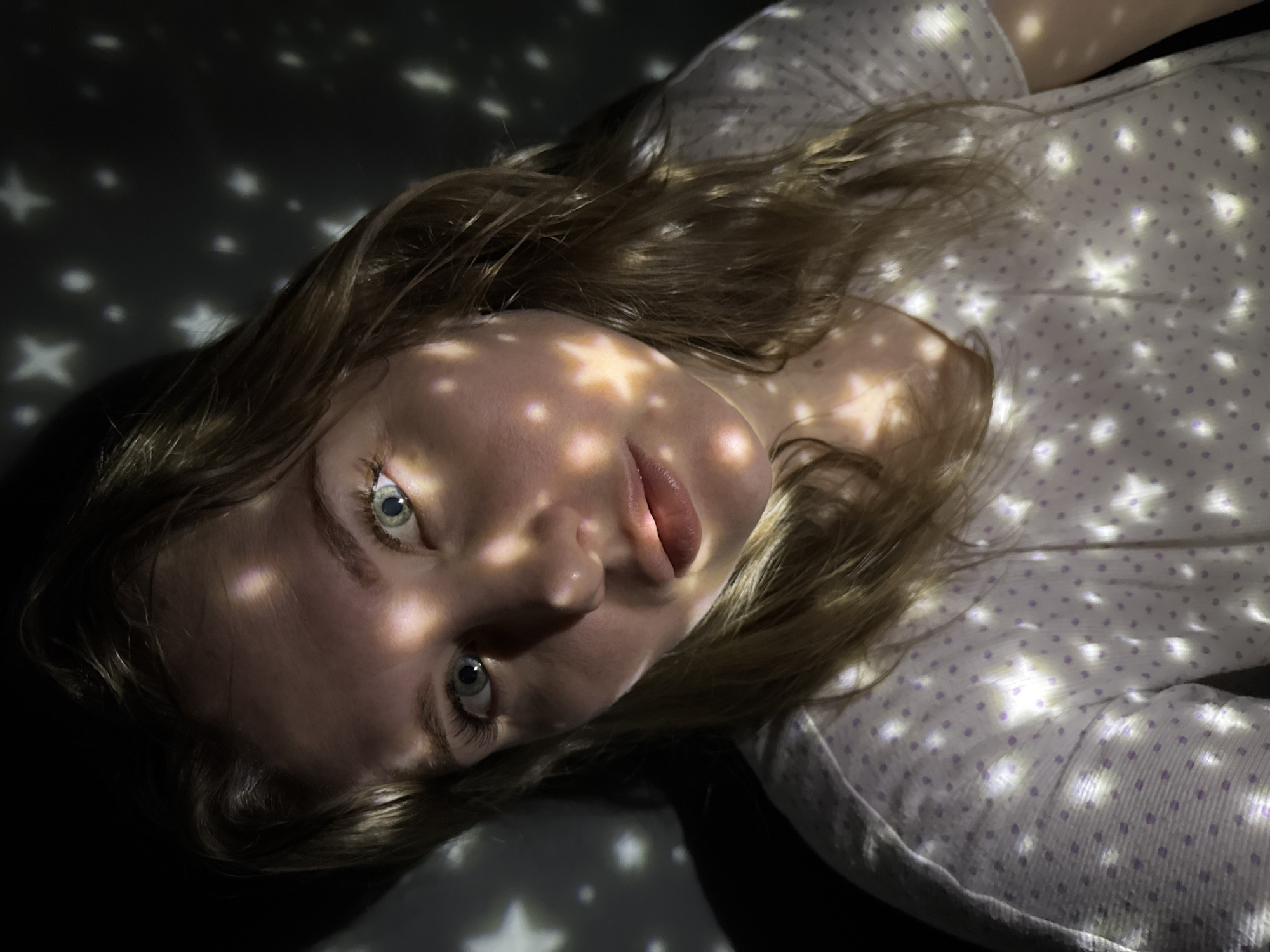 A close-up, high-angle portrait captures a young adult female in an indoor, artificially lit environment, specified as being in the city of Gornyi (Dvurechenskoe S/P), Russia. The subject possesses fair skin, light brown hair, and light blue-green eyes, maintaining a direct, neutral gaze toward the camera. She is clad in a white, short-sleeved shirt featuring a subtle pattern of light purple polka dots. The background is dark and unidentifiable. Both the subject and the surrounding dark space are illuminated by numerous projected white light patterns, consisting of distinct stars and smaller circular dots, which vary in size and intensity. These light patterns create highlights across her face, hair, and clothing, as well as on the dark surfaces behind her.