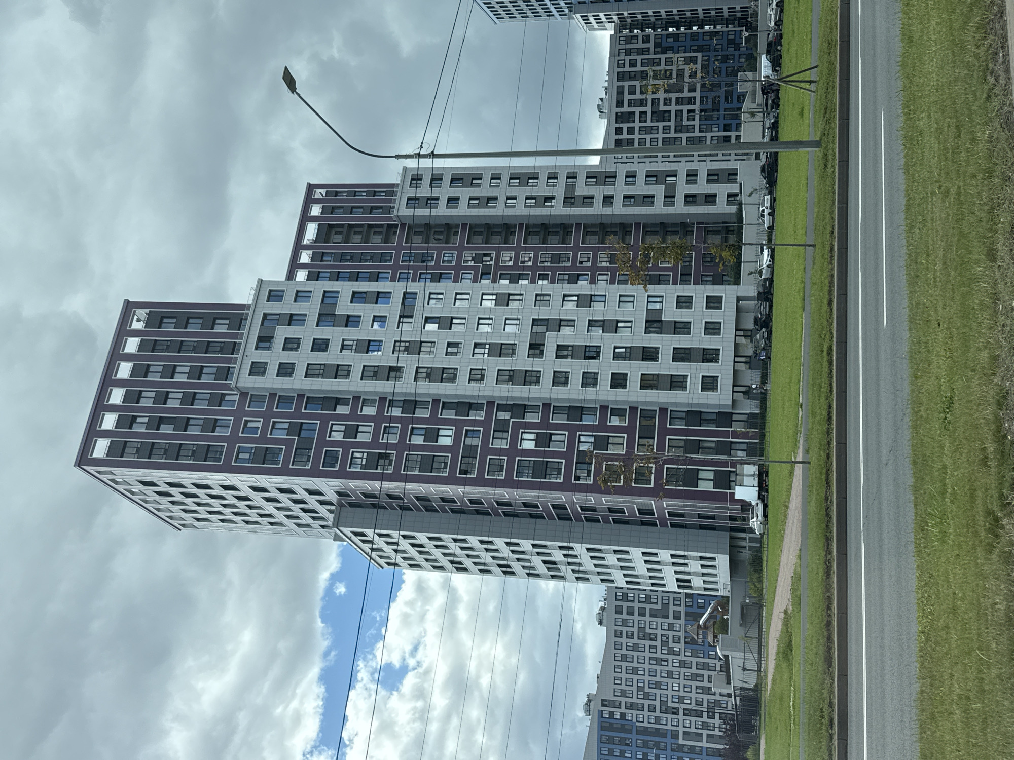 The image depicts a street view in Saint Petersburg, Russia, focusing on modern residential buildings. In the foreground, a wide asphalt road with white lane markings runs horizontally, bordered by a strip of green grass and a curb. Behind the road, a manicured green lawn stretches towards a pathway. A row of parked cars and a delivery truck are visible on the lawn near a fence. Several multi-story buildings with contemporary architecture, featuring a mix of light grey and dark purple facades with numerous windows, dominate the background. The sky is overcast with patches of blue sky and white clouds. A street lamp with a curved arm is positioned to the right of the frame, and power lines crisscross the upper portion of the image.