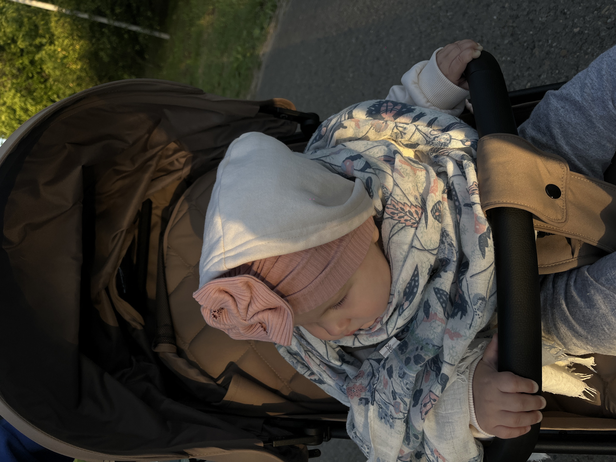 A baby is nestled in a stroller. The baby wears a pink hat adorned with a flower and a white hooded sweatshirt. A patterned scarf is draped around the baby. The stroller is on an asphalt surface. Green foliage is visible in the background. The location is Novopokrovskaya, Russia.