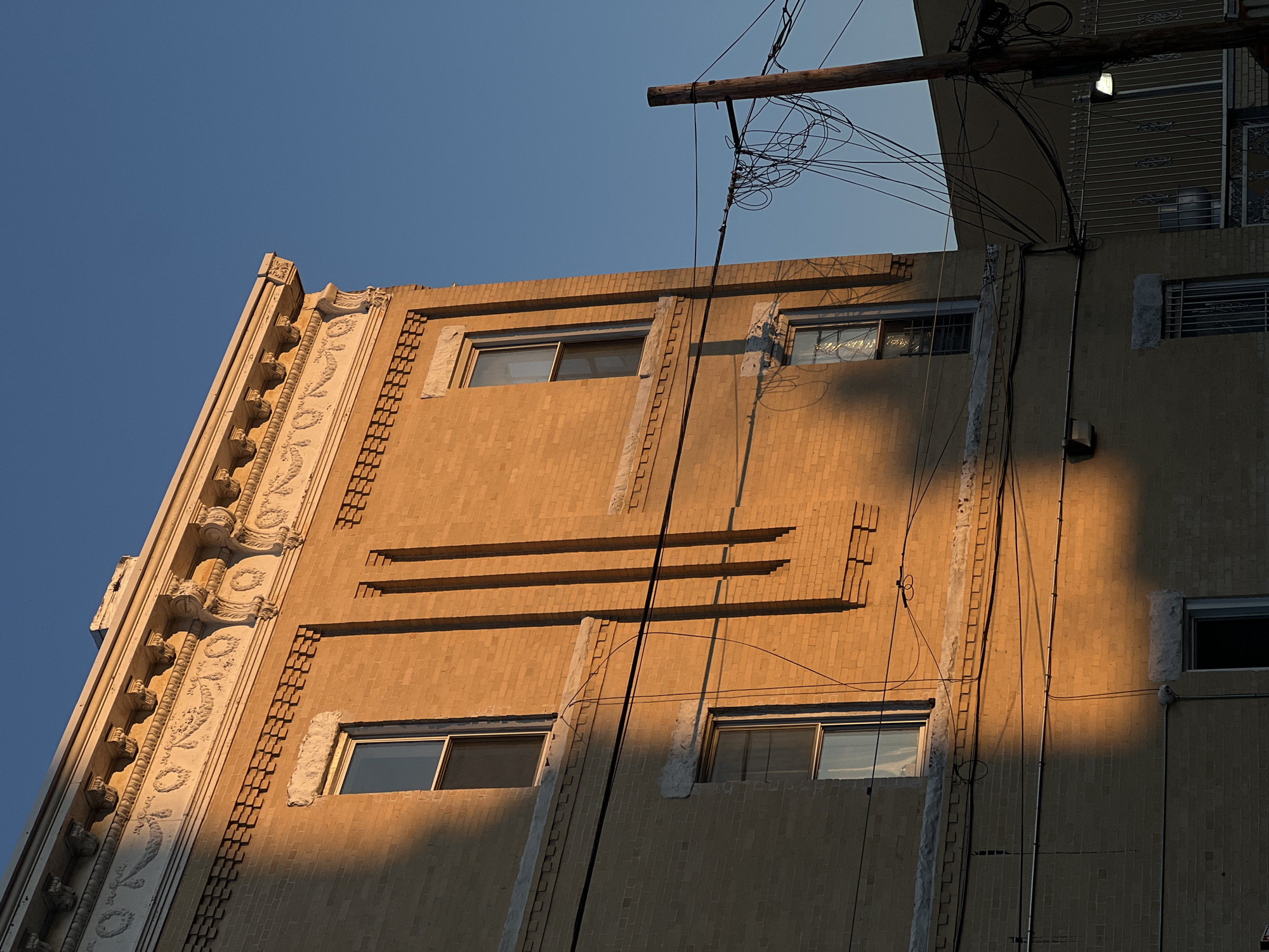 The photograph captures a section of a building facade under a clear, blue sky. The architecture of the building exhibits a combination of brickwork and decorative elements, with a vertical row of raised embellishments running along the left side. The building has rectangular windows with visible frames. Horizontal lines are incorporated into the brick pattern on the face of the building.
Power lines and associated poles are visible in the upper right portion of the image, creating a network of wires against the sky. The lighting indicates it may be late afternoon or early morning. Shadows are cast on the building, creating gradients in the color tones and texture. The location is potentially within the city of New York, United States of America. There are no people visible in the image.
