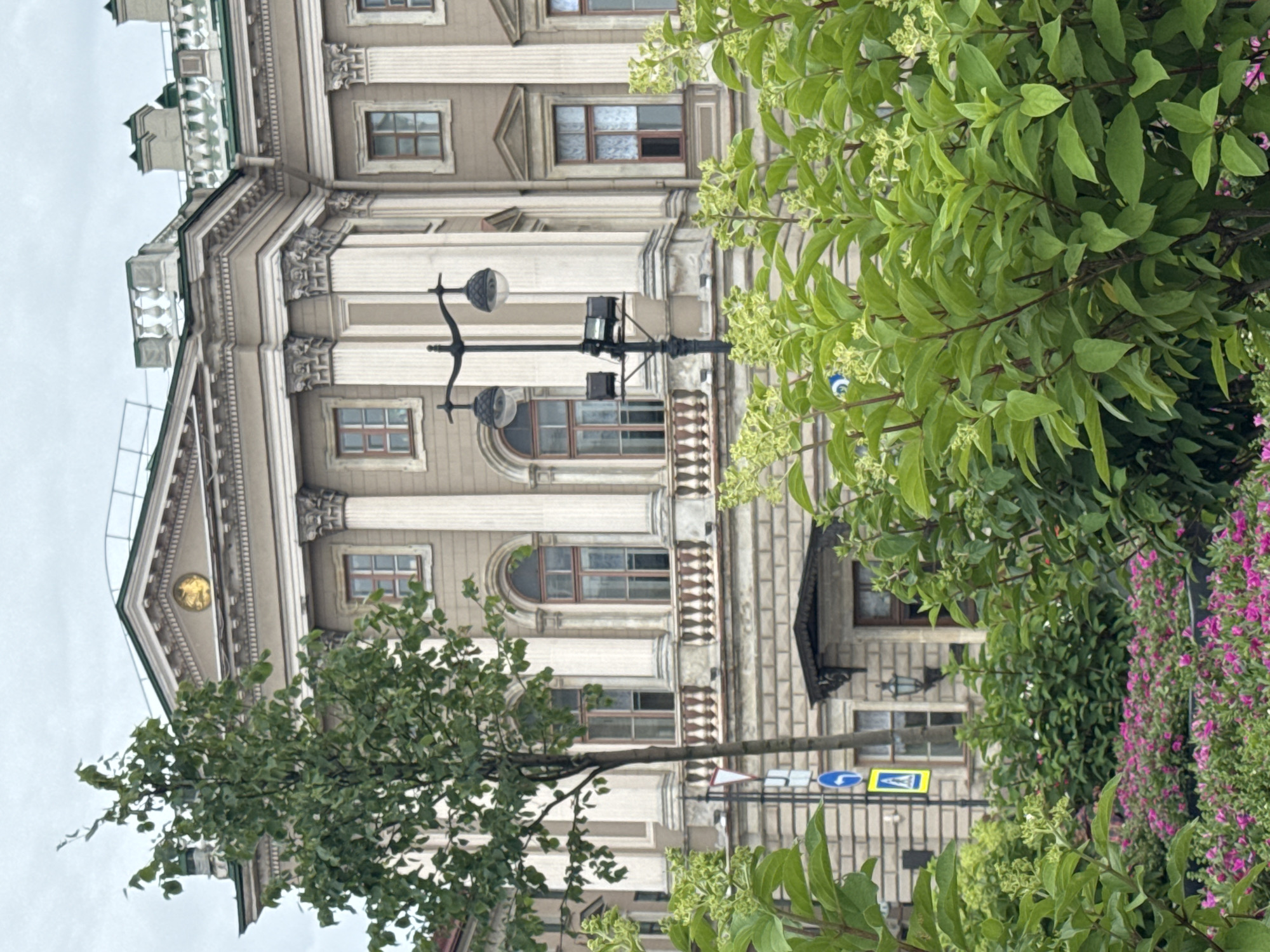 The image shows the facade of a large, classical building, likely a museum or government institution, in Saint Petersburg, Russia. The building has a beige-colored stone facade with numerous columns, arched windows, and ornate detailing. In the foreground, lush green foliage and pink flowers obscure the lower part of the building. A street lamp with two globe-shaped lights and additional lighting fixtures stands to the left of the frame. Several traffic signs are visible behind the foliage, including a yield sign and a blue circular sign with a white arrow indicating a left turn. A single tree with green leaves stands to the left of the building, partially obscuring the facade. The sky is overcast and grey, suggesting a cloudy day. The overall impression is one of historical grandeur and urban greenery.