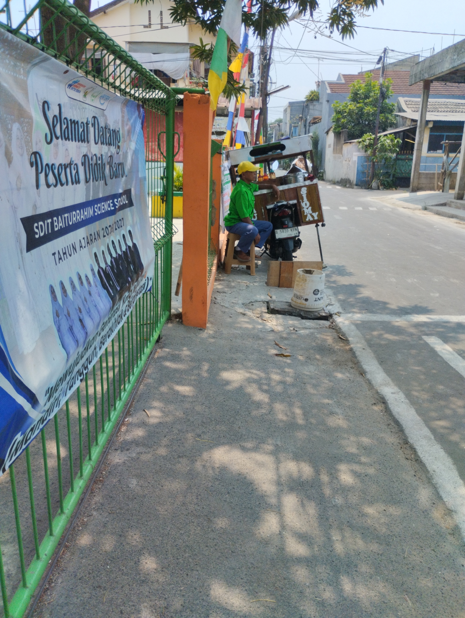 In Bekasi, Indonesia, on a sunny day, a street vendor sits on a stool next to their food cart, which is attached to a motorcycle. The cart displays the word "SIOMAY" prominently. The vendor, wearing a green shirt and a yellow cap, appears to be taking a break. The scene is located alongside a paved road with a white line marking its edge. In the background, residential buildings with tiled roofs are visible. To the left of the vendor, a green metal fence borders a sidewalk, and a banner with Indonesian text, likely a school welcome sign, is attached to it. The ground is a mixture of asphalt and dirt, with some scattered leaves and debris. The overall impression is of a typical urban street scene in Indonesia.