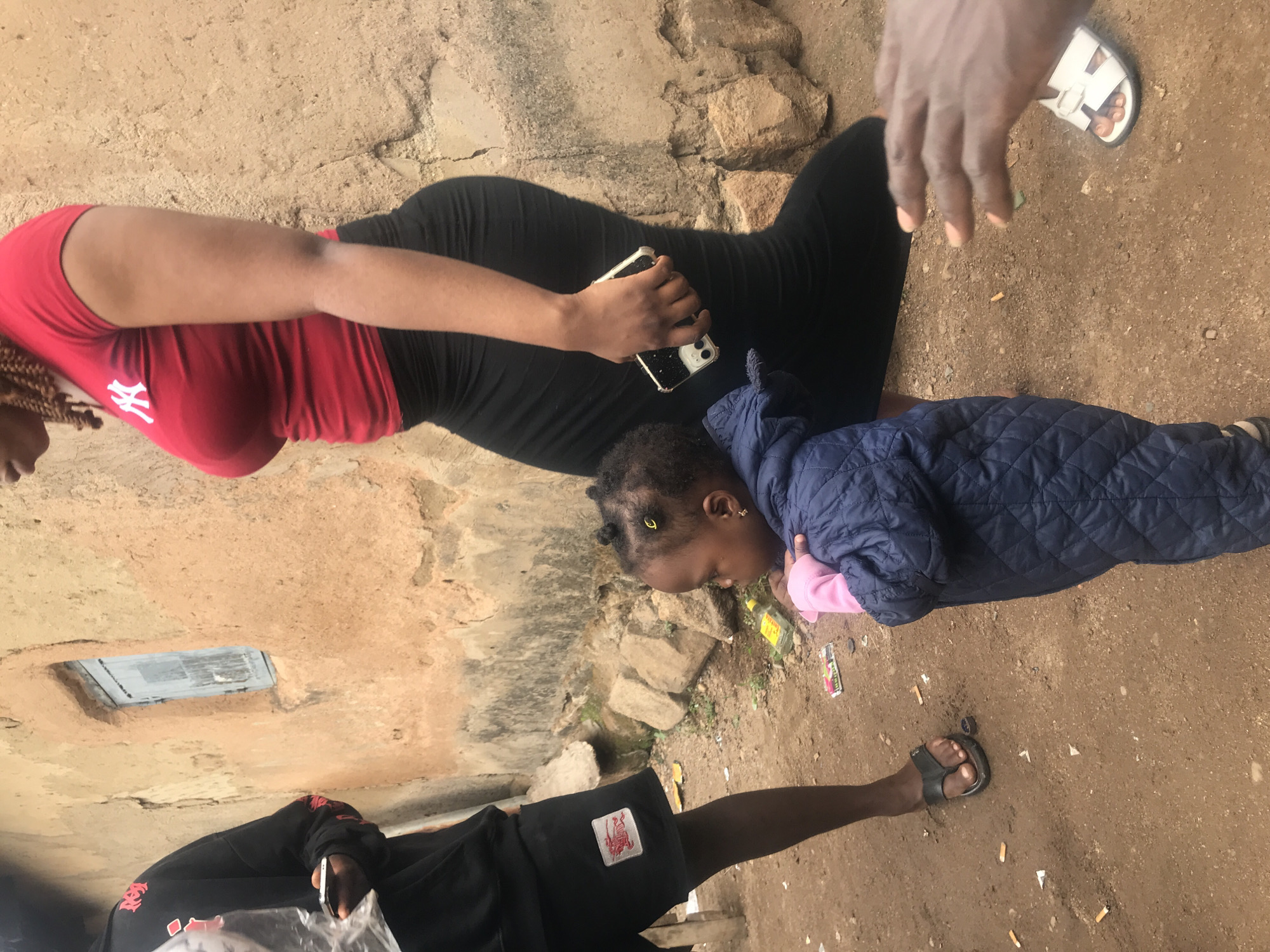 A group of people, including a woman in a red t-shirt and black skirt, a toddler in a blue jacket, and a partial view of another person, are gathered on a dirt path outside a mud-brick building in Kabong, Nigeria. The building has a small, weathered blue window. The ground is littered with cigarette butts and small pieces of paper. The woman in the red shirt holds a smartphone. The toddler is looking down and appears to be holding something pink. The other person visible is wearing a black hoodie and black shorts with a red logo.