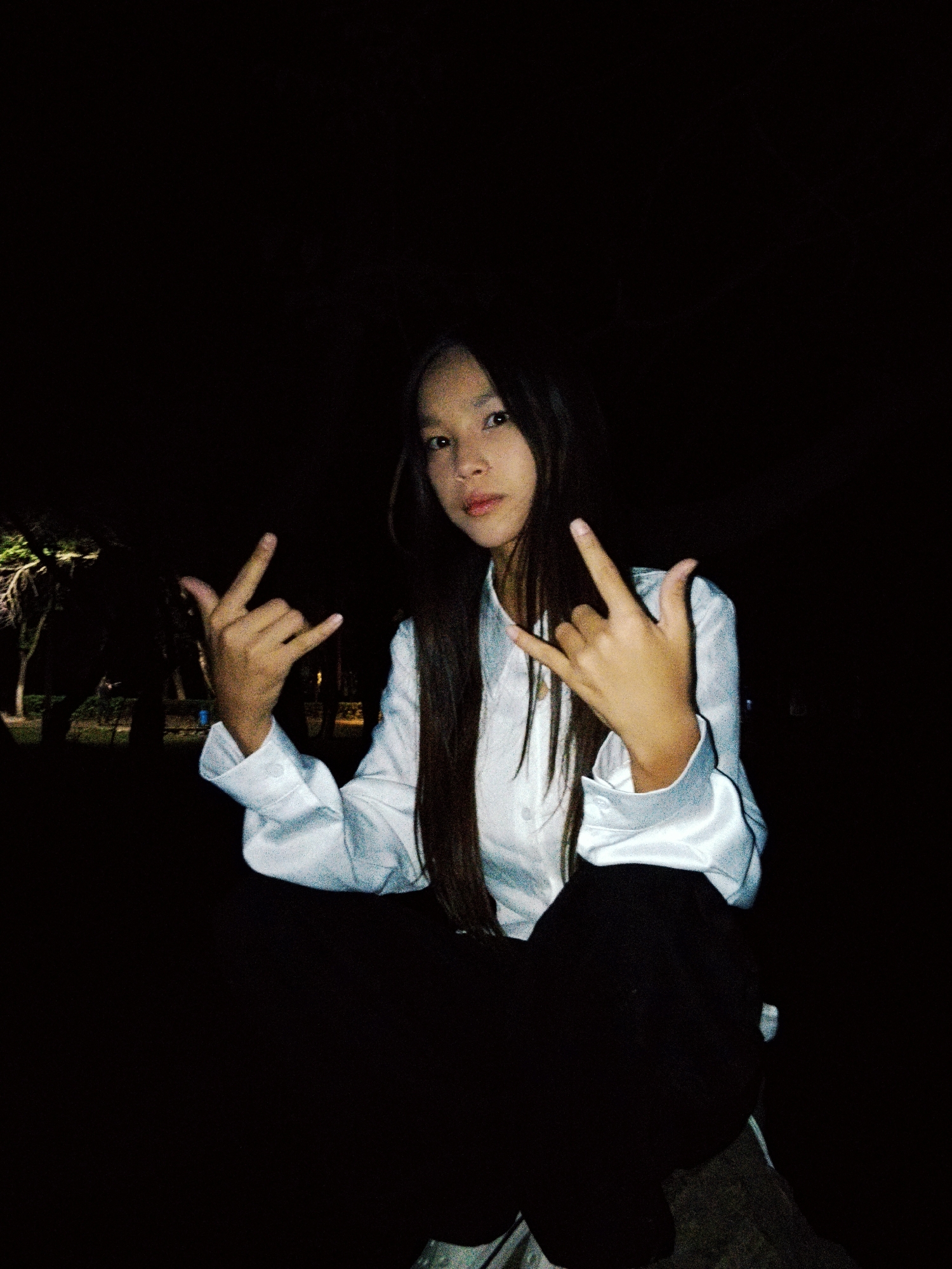 In a dimly lit outdoor setting, a young person with long, dark hair is seated and looking at the camera. They are wearing a white collared shirt and dark pants. Their hands are raised, and they are making a hand gesture with both index fingers and thumbs extended, resembling the "rock on" sign. The background is dark with some indistinct shapes of trees and foliage visible on the left. The scene appears to be in an urban park or public space within Bishkek City, Kyrgyzstan, during nighttime.
